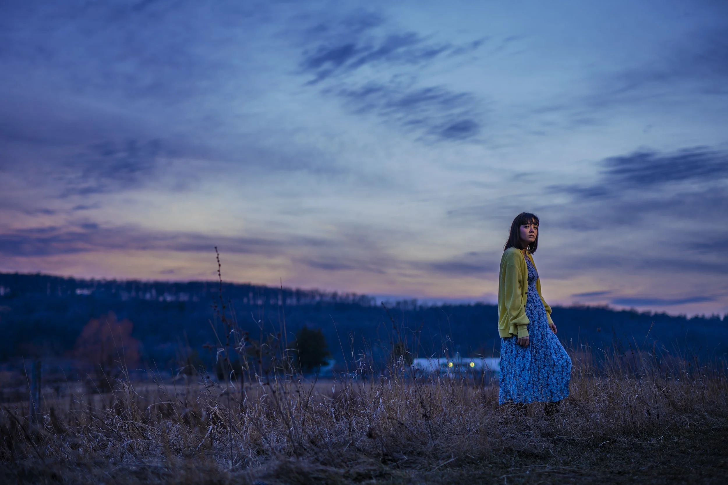 A young woman standing alone in a field during dusk, with a sky painted in shades of blue and purple, wearing a yellow jacket and a blue floral dress.