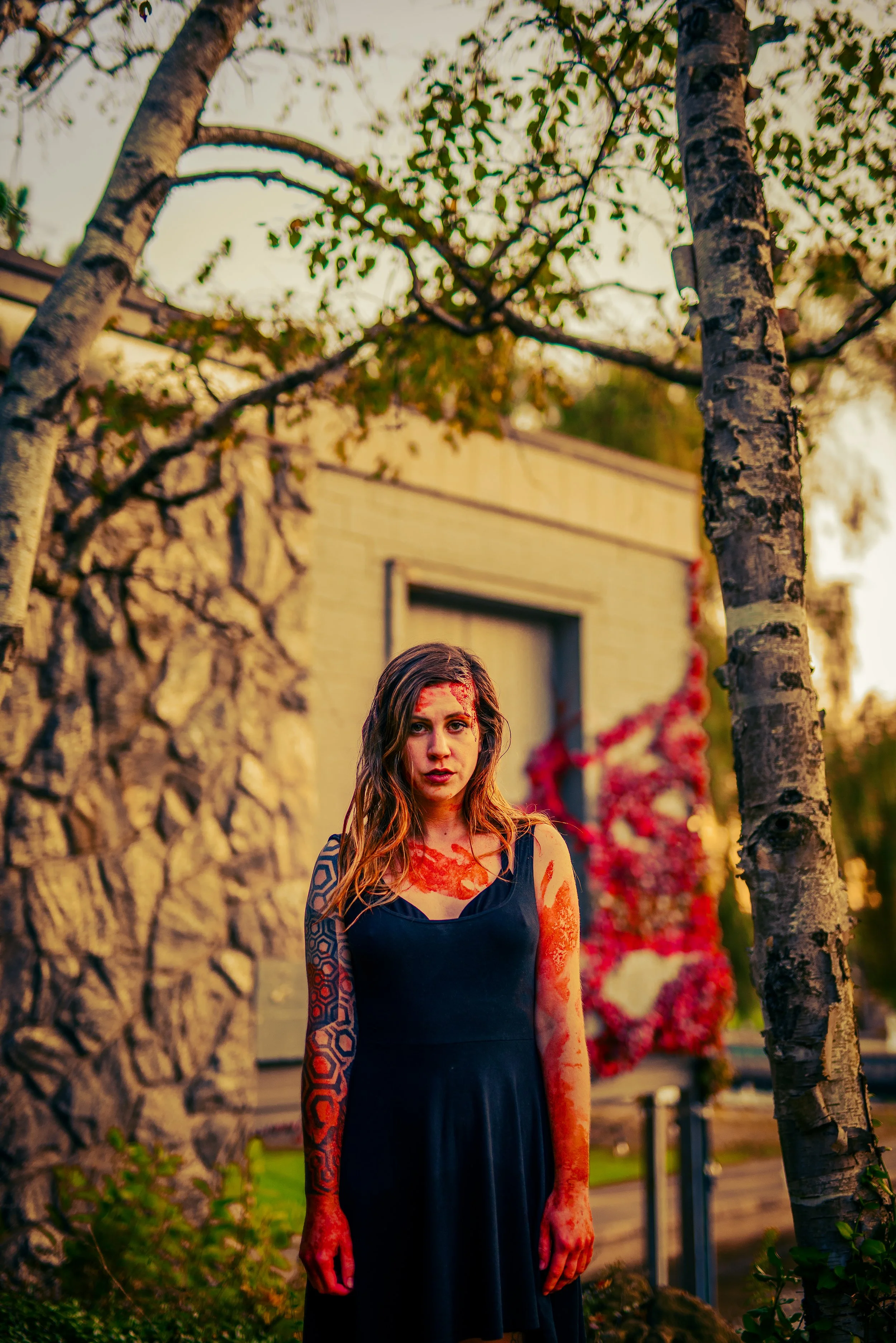 A woman with long wavy brown hair and blue eyes stands outdoors among trees, wearing a black sleeveless dress. Her arms and parts of her face are covered in red paint or blood, creating a horror or Halloween-themed appearance. In the background, ther