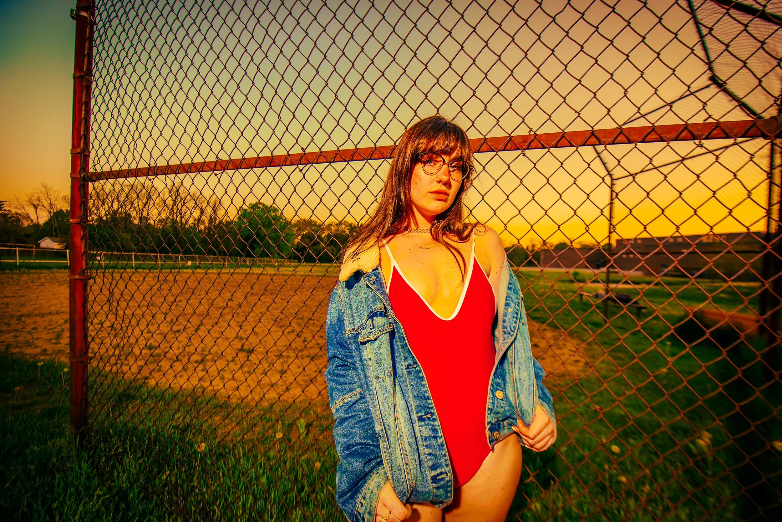 A young woman with long brown hair, glasses, and a septum piercing, wearing a red swimsuit, denim jacket, and a choker, standing in front of a chain-link fence during sunset.