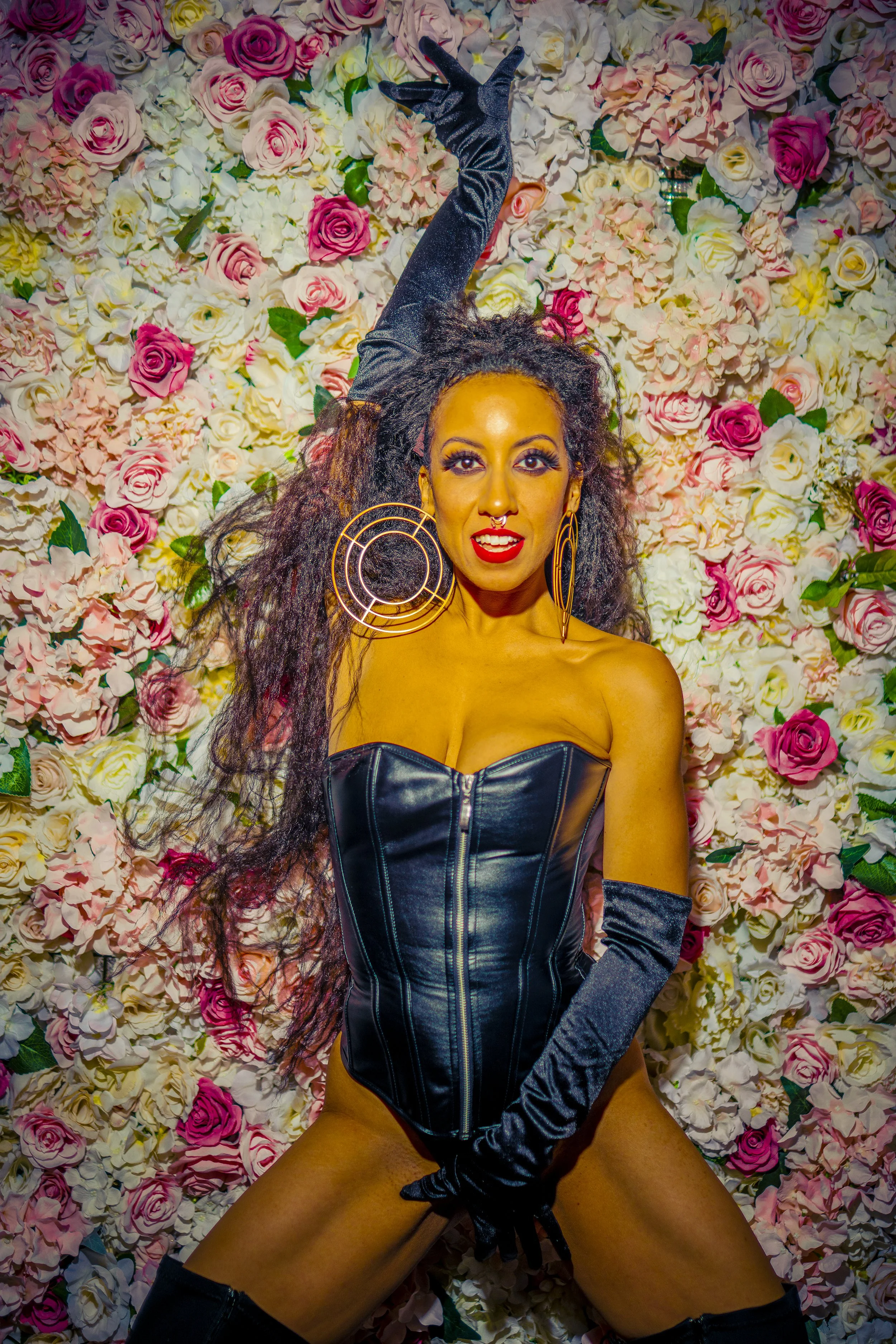 A woman with curly hair and large hoop earrings posing in front of a floral wall, wearing a black leather corset, long black gloves, and fishnet stockings.