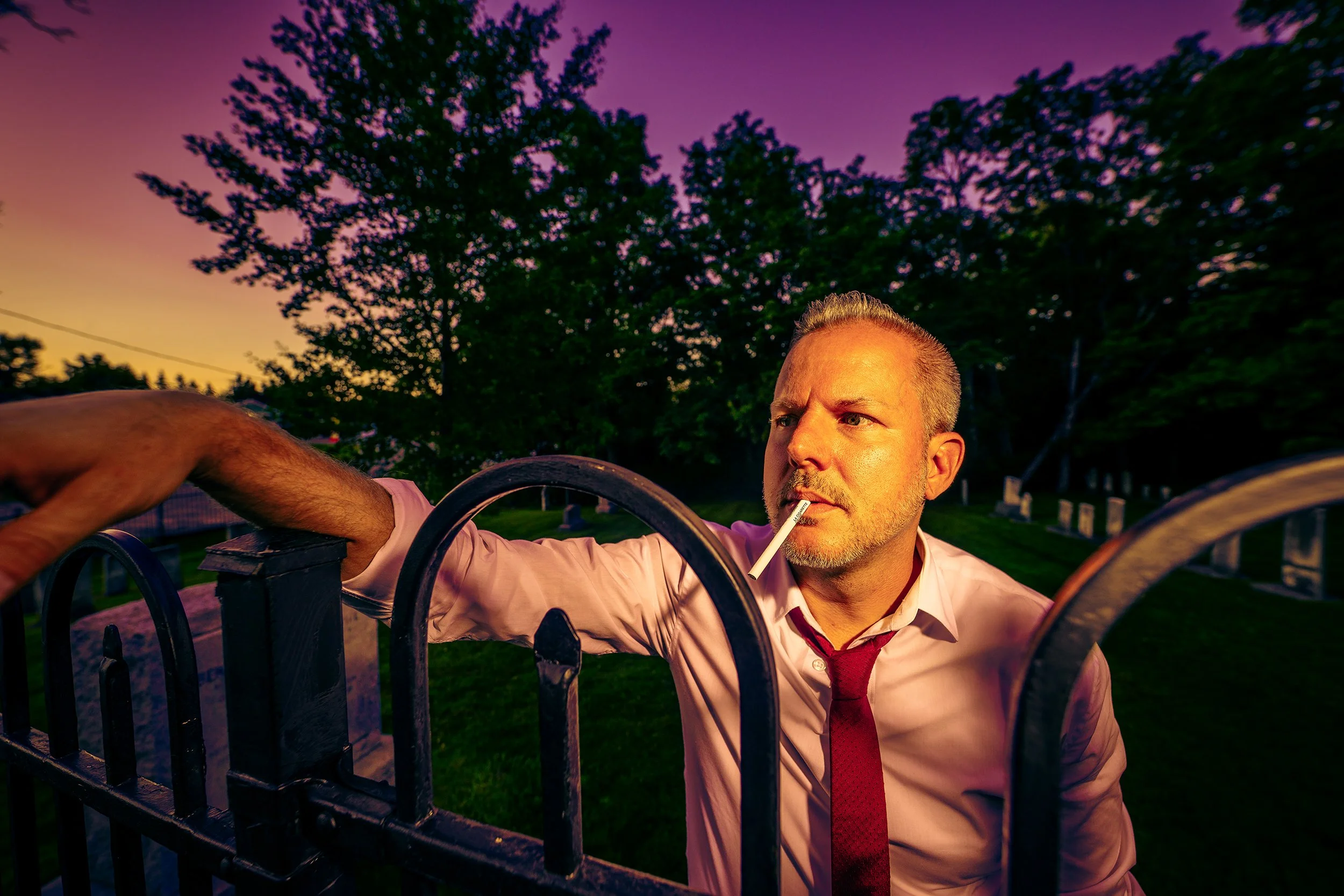 A man with gray hair and a serious expression, wearing a white shirt and red tie, is leaning on a black metal fence at sunset. He is holding a cigarette in his mouth. The background features trees and a colorful sky with shades of purple, pink, and y