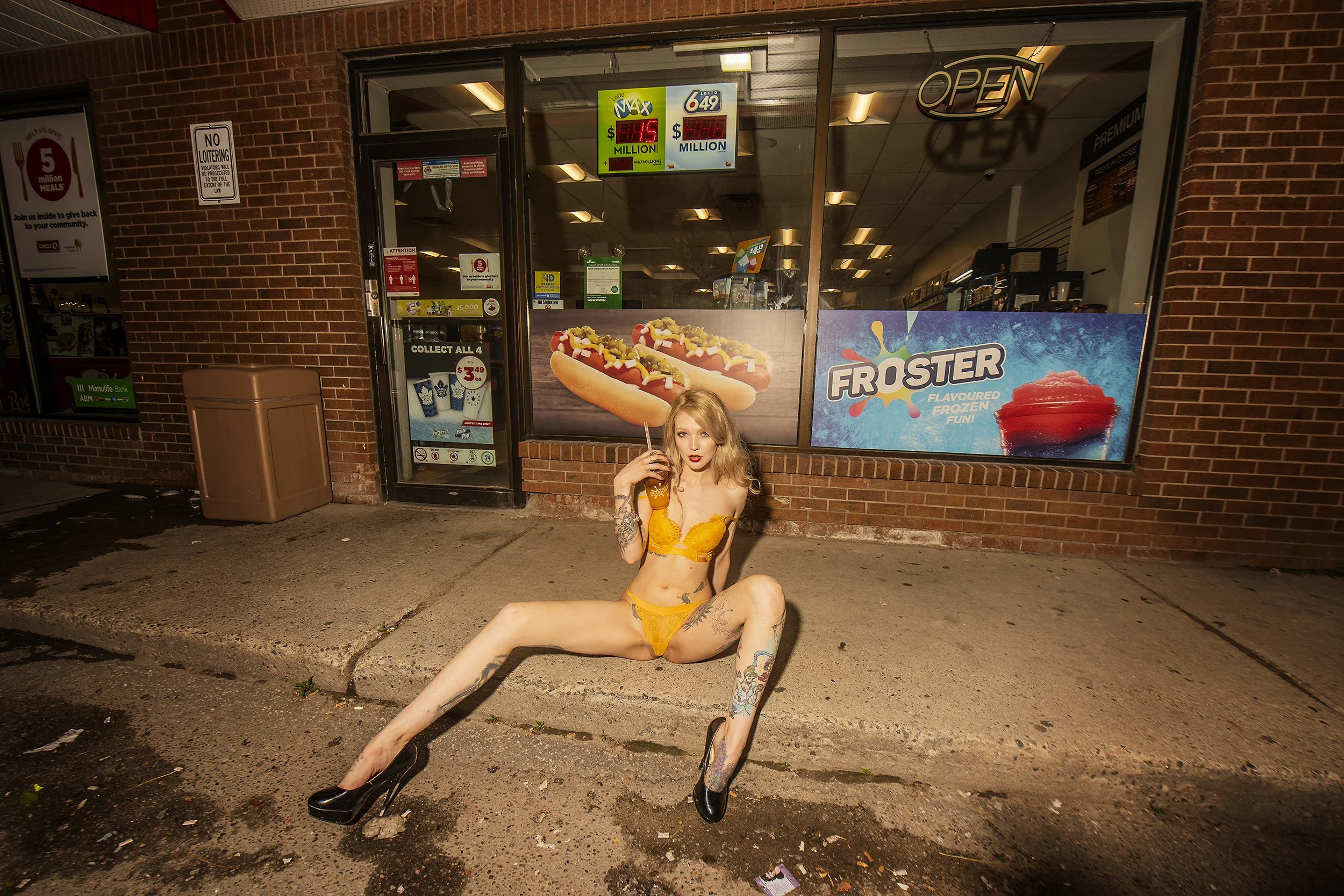 A woman wearing an orange lingerie set and black high heels sitting on the sidewalk outside a fast food restaurant, holding a drink and posing with her legs spread apart.