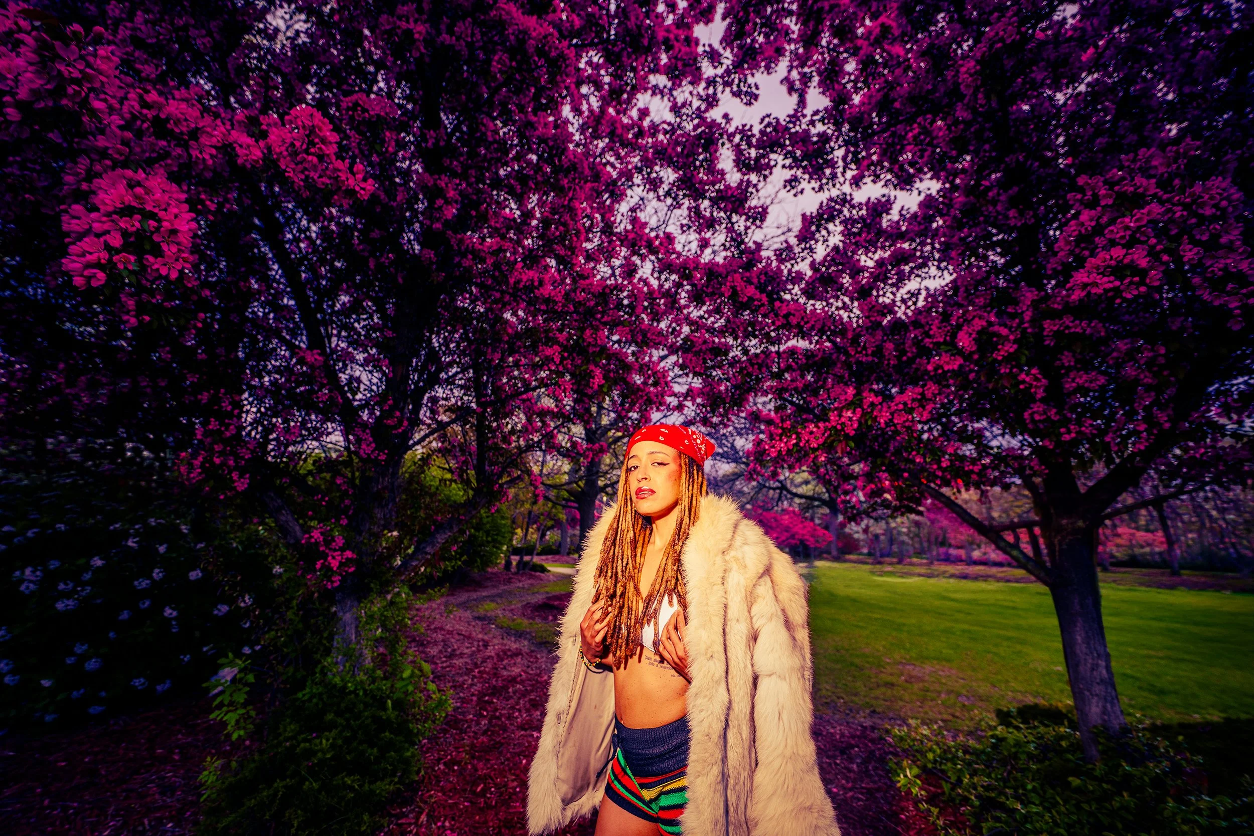 A woman with dreadlocks wearing a red bandana, beige fur coat, colorful shorts, and a beige crop top standing outdoors under blooming pink and purple flowering trees.