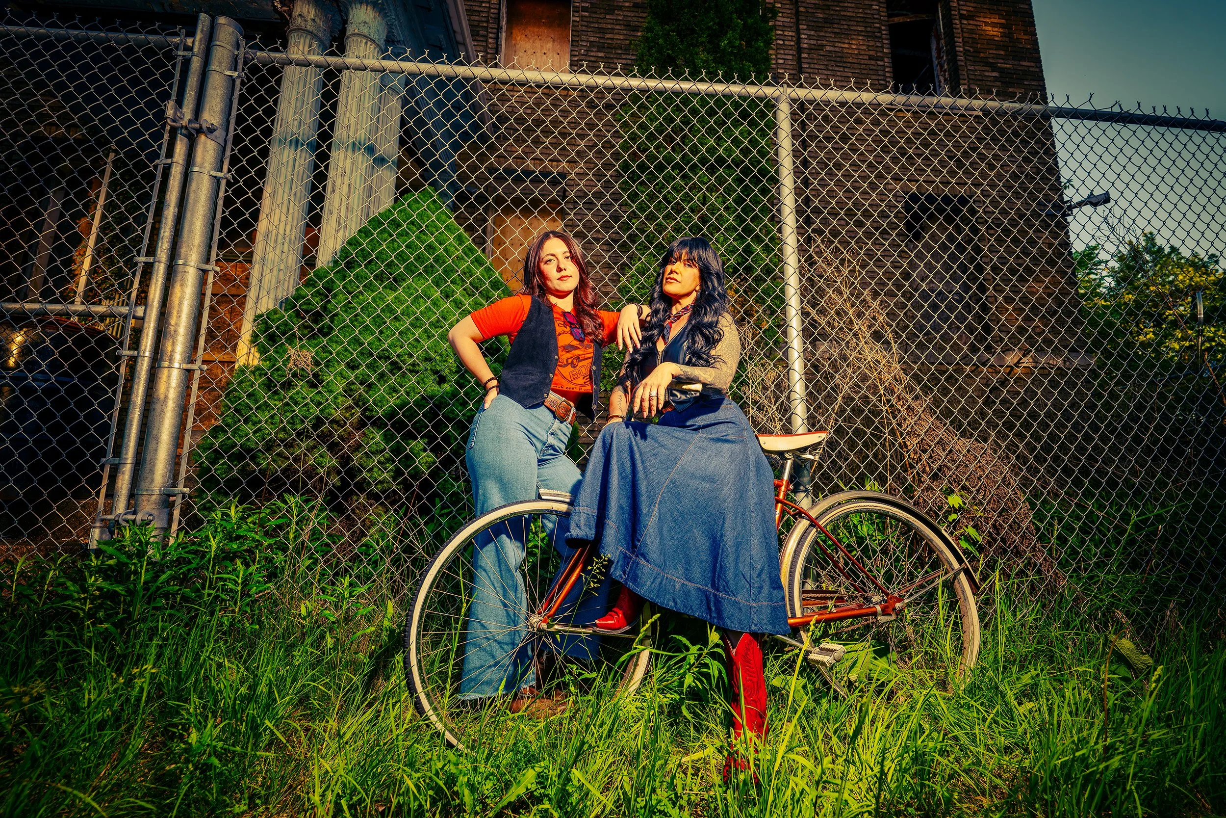Two women in front of a chain-link fence, one standing and the other sitting on a bicycle, with a dilapidated brick building in the background.