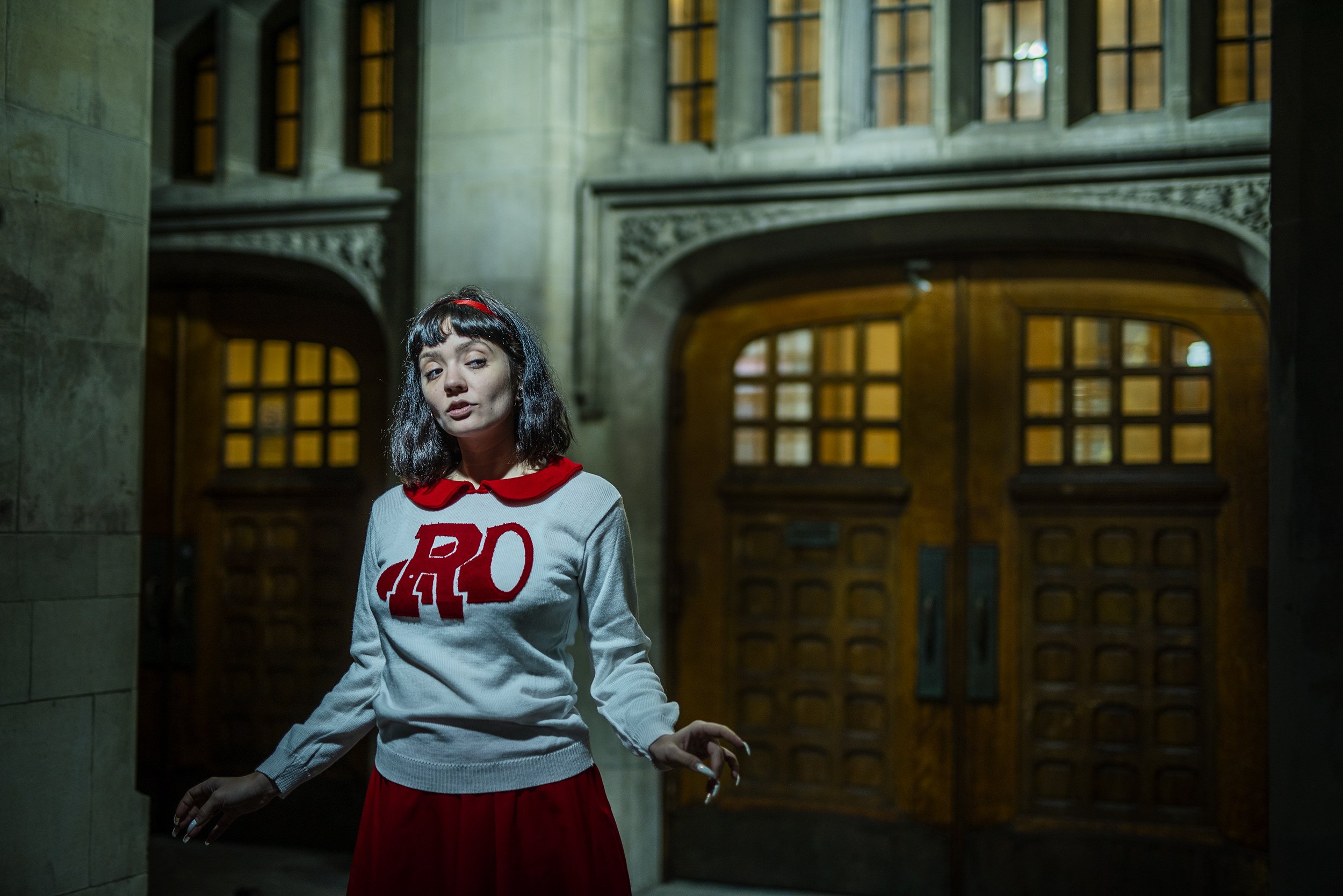 A young woman with black hair, wearing a gray sweatshirt with red letters and a red skirt, stands inside a large building with high ceilings, arched wooden doors, and stained glass windows.