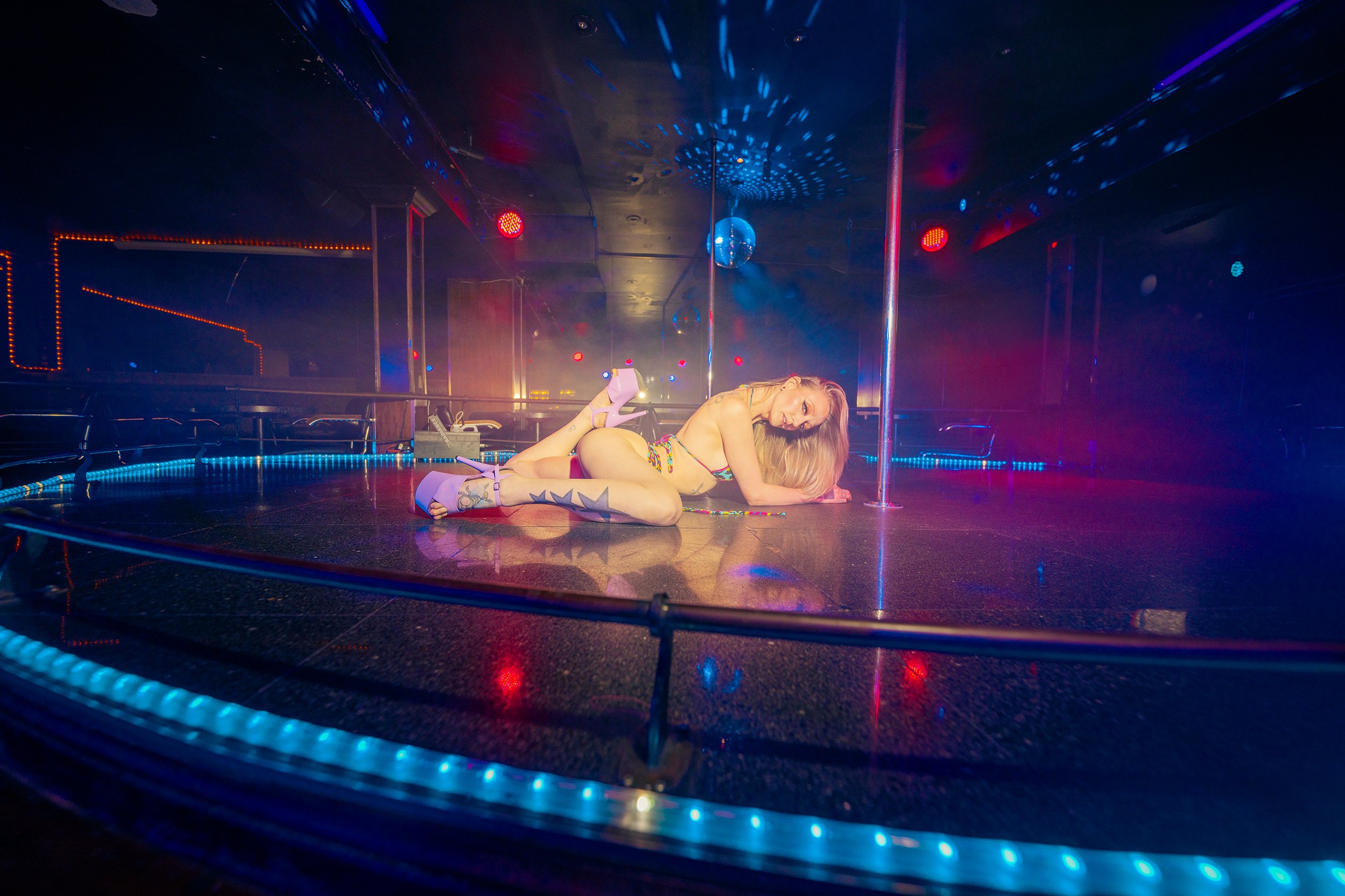 A woman with tattoos on her left leg posing on a dance pole in a dimly lit club, illuminated by colorful lights.