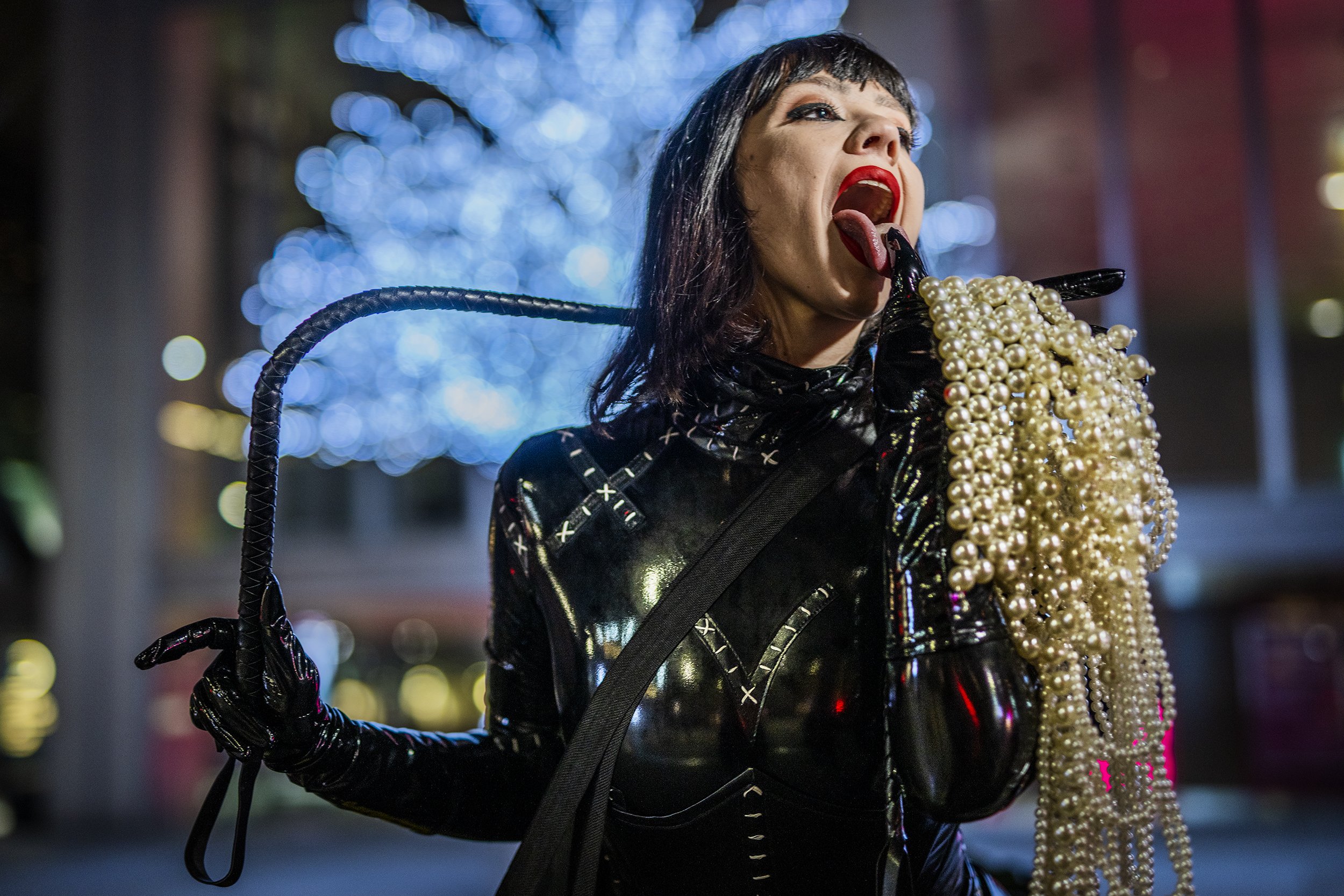 A woman with black hair and red lipstick is holding a pearl necklace to her mouth, wearing black gloves and a shiny black outfit, against a background of blue and white lights.