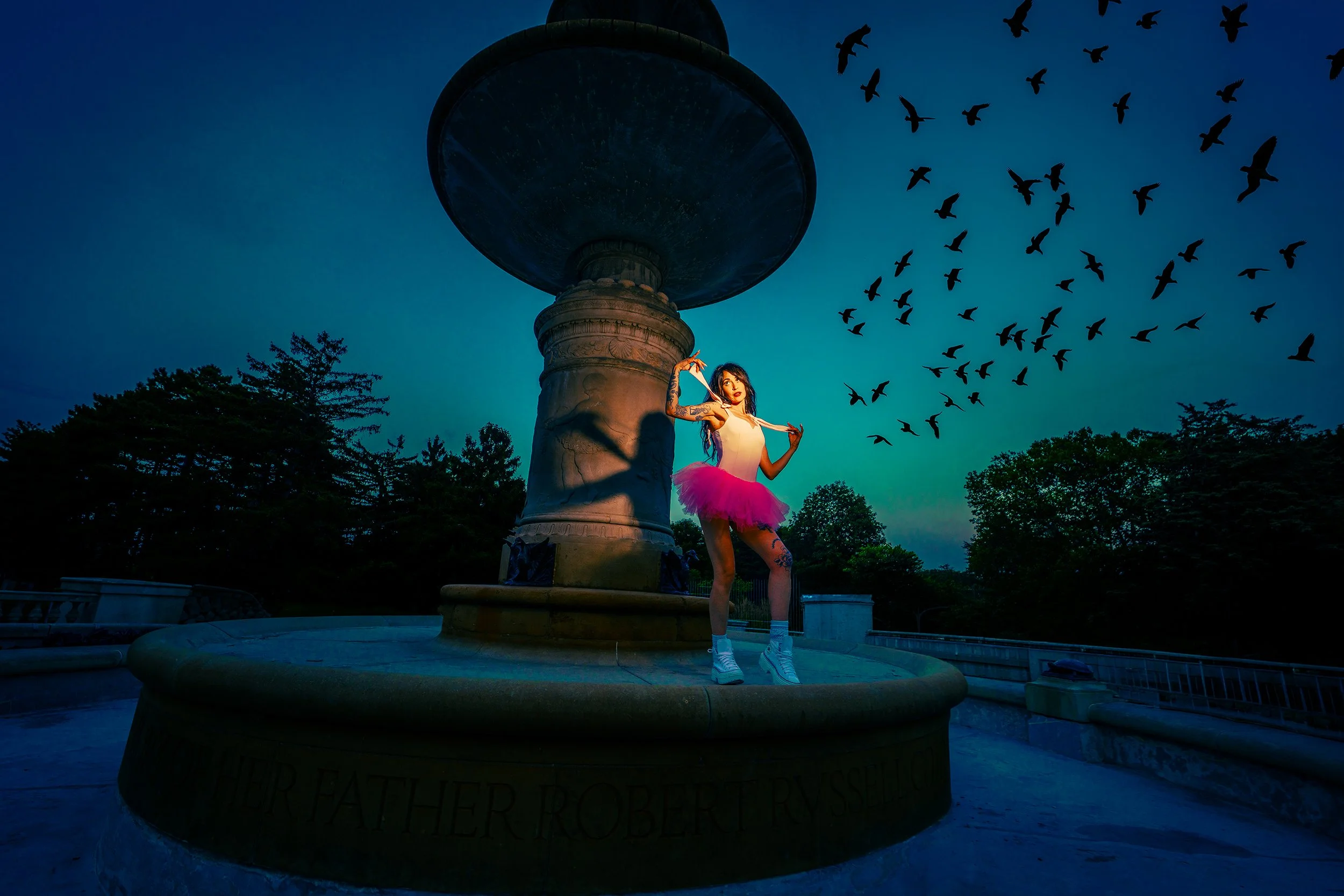 A woman in a pink tutu and white top standing in front of a large fountain with a bird-shaped ornament on top, with a flock of birds flying in the evening sky.