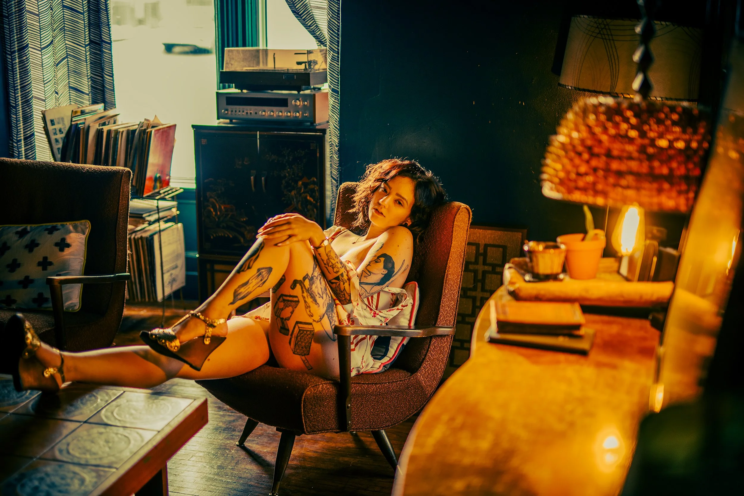 A woman with tattoos lounging in a vintage-style room with colorful lighting, surrounded by books, vinyl records, and a record player.