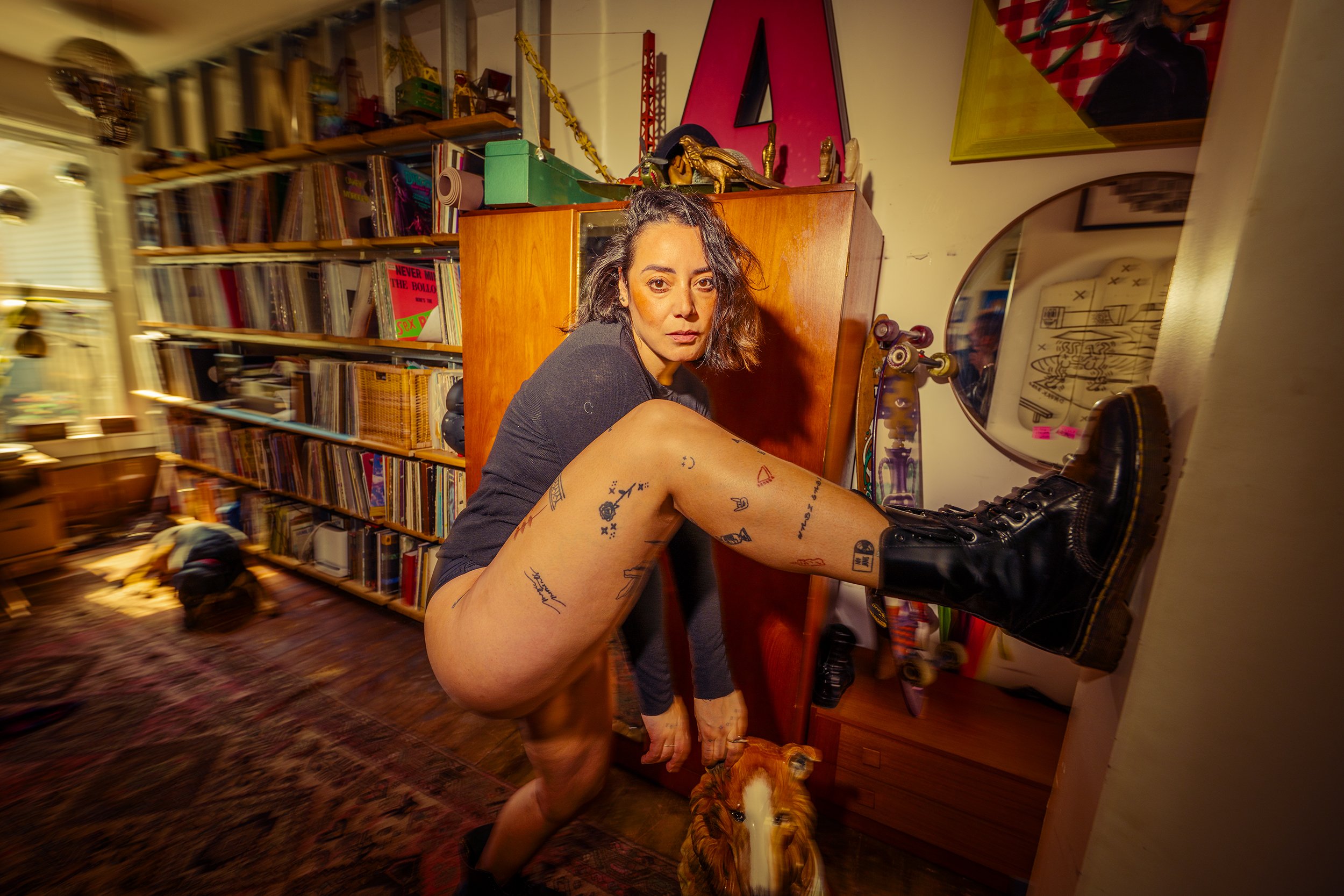 A woman with tattoos on her legs, wearing combat boots, crouching indoors with one leg raised on a wall, in a cluttered room filled with books, skateboards, and art. She has dark, shoulder-length hair and looks directly at the camera.