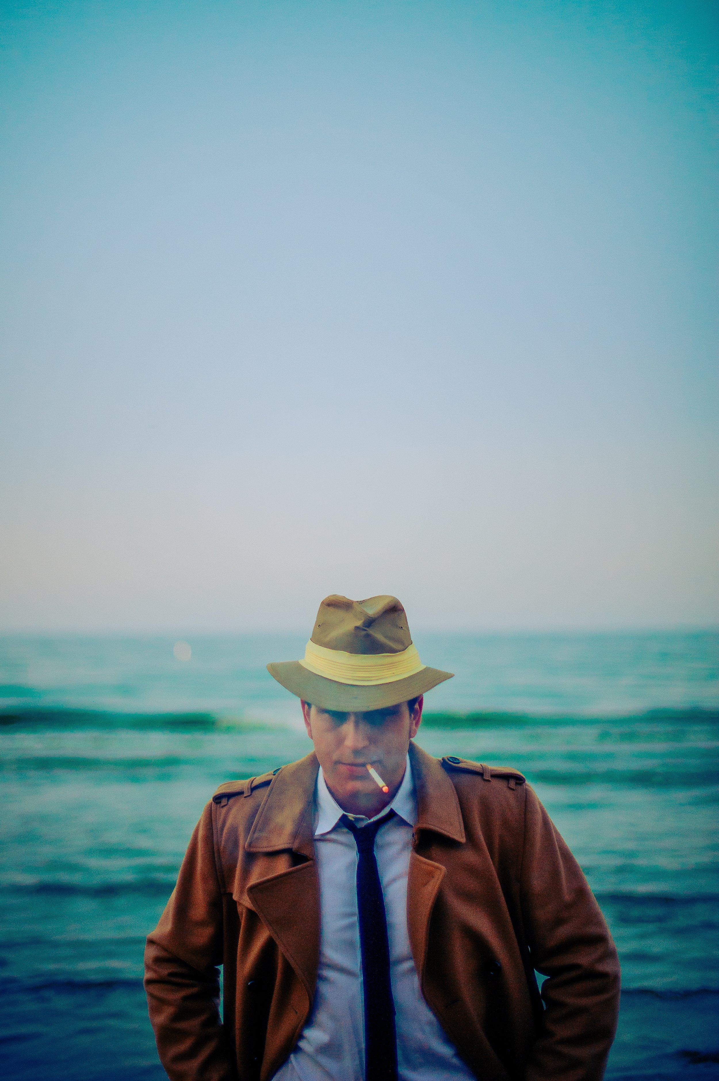 A man wearing a brown trench coat, white shirt, and blue tie standing in front of the ocean with a hat and a lit cigarette in his mouth.