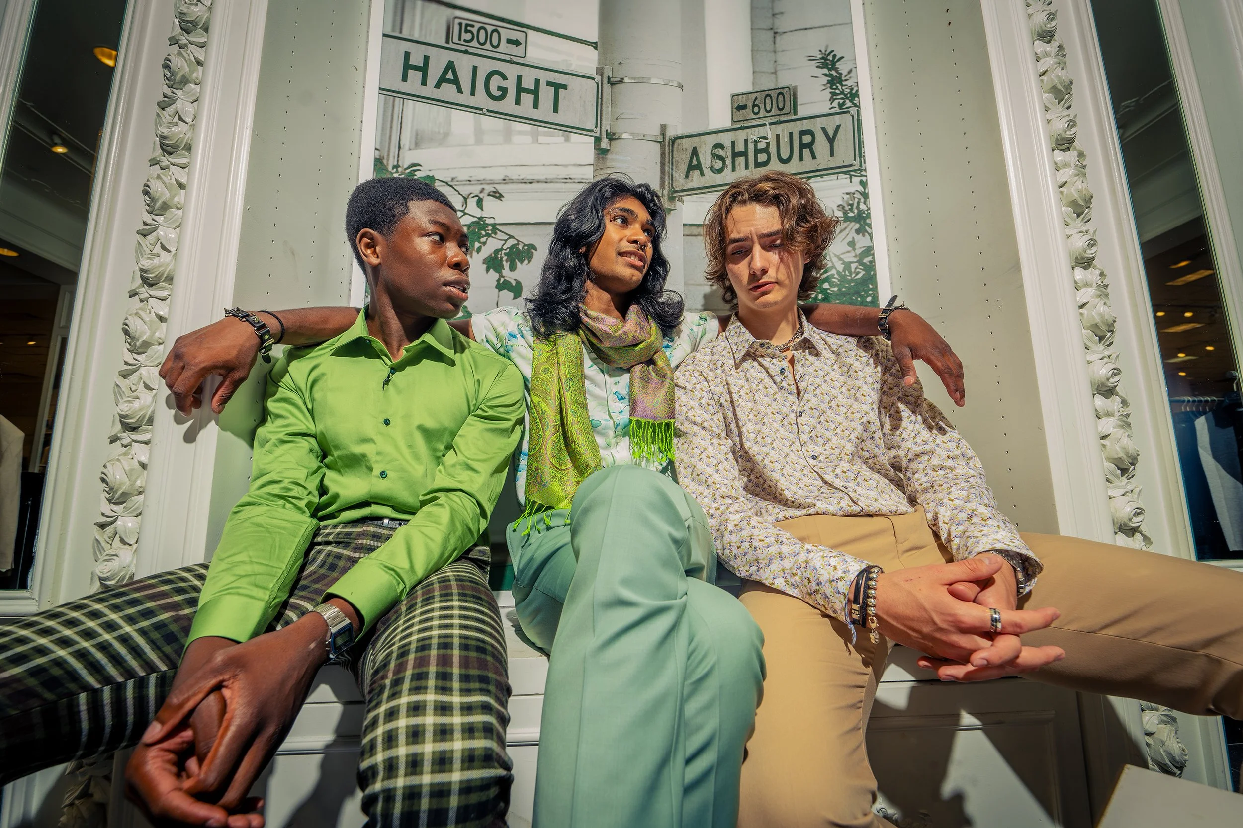 Three young adults sitting closely together with arms around each other, inside a decorated space with street signs reading 'Haight' and 'Ashbury' behind them.