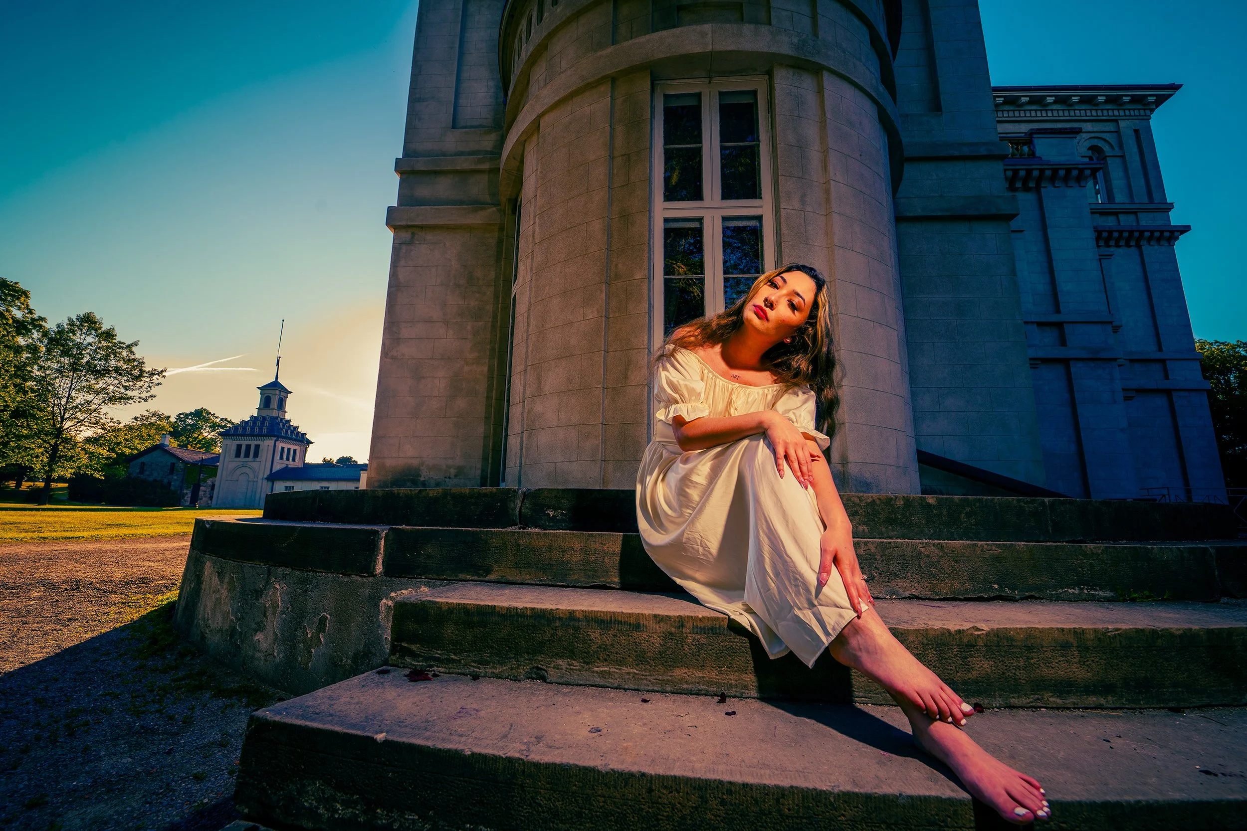 A woman in a cream-colored dress sitting on the steps in front of historic buildings during sunset. She has long, wavy hair and is posing with a serious expression while looking at the camera.