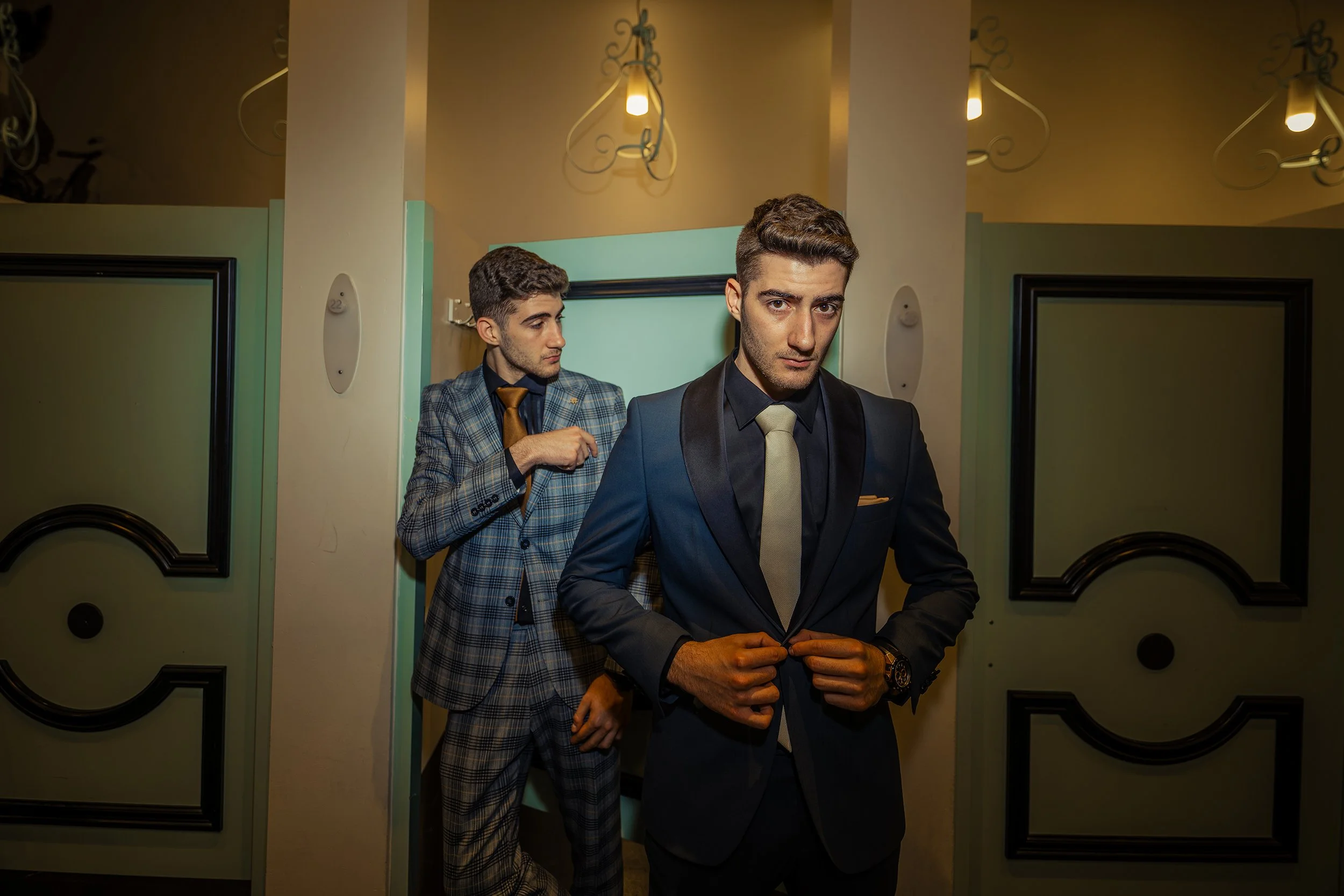 Two men dressed in suits, one adjusting his cufflinks, standing in an interior hallway with pastel-colored walls and decorative lighting fixtures.