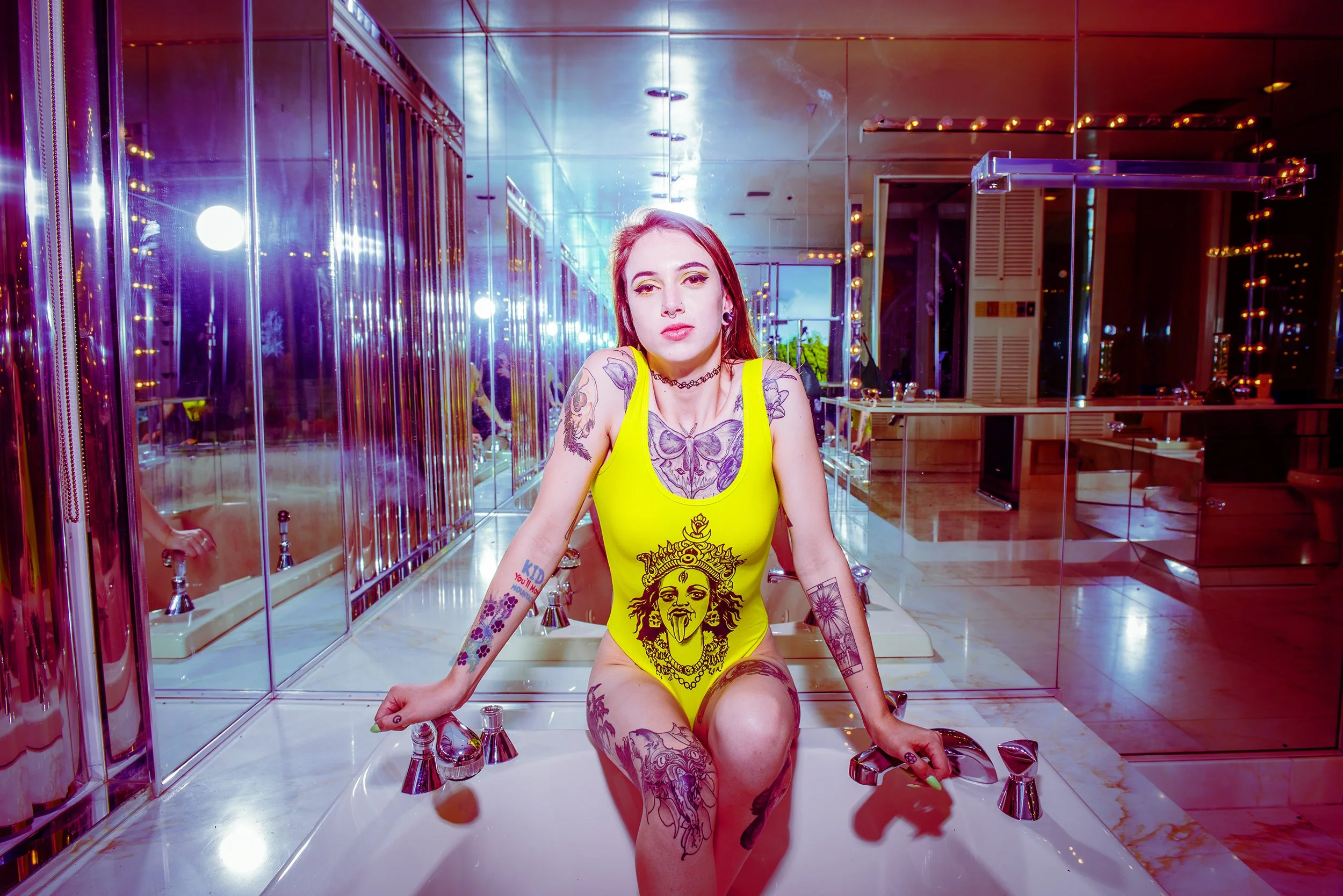 A woman with tattoos and piercings wearing a yellow tank top with a graphic design, sitting in a marble sink with multiple faucets in a room with mirrored walls and colorful lighting.
