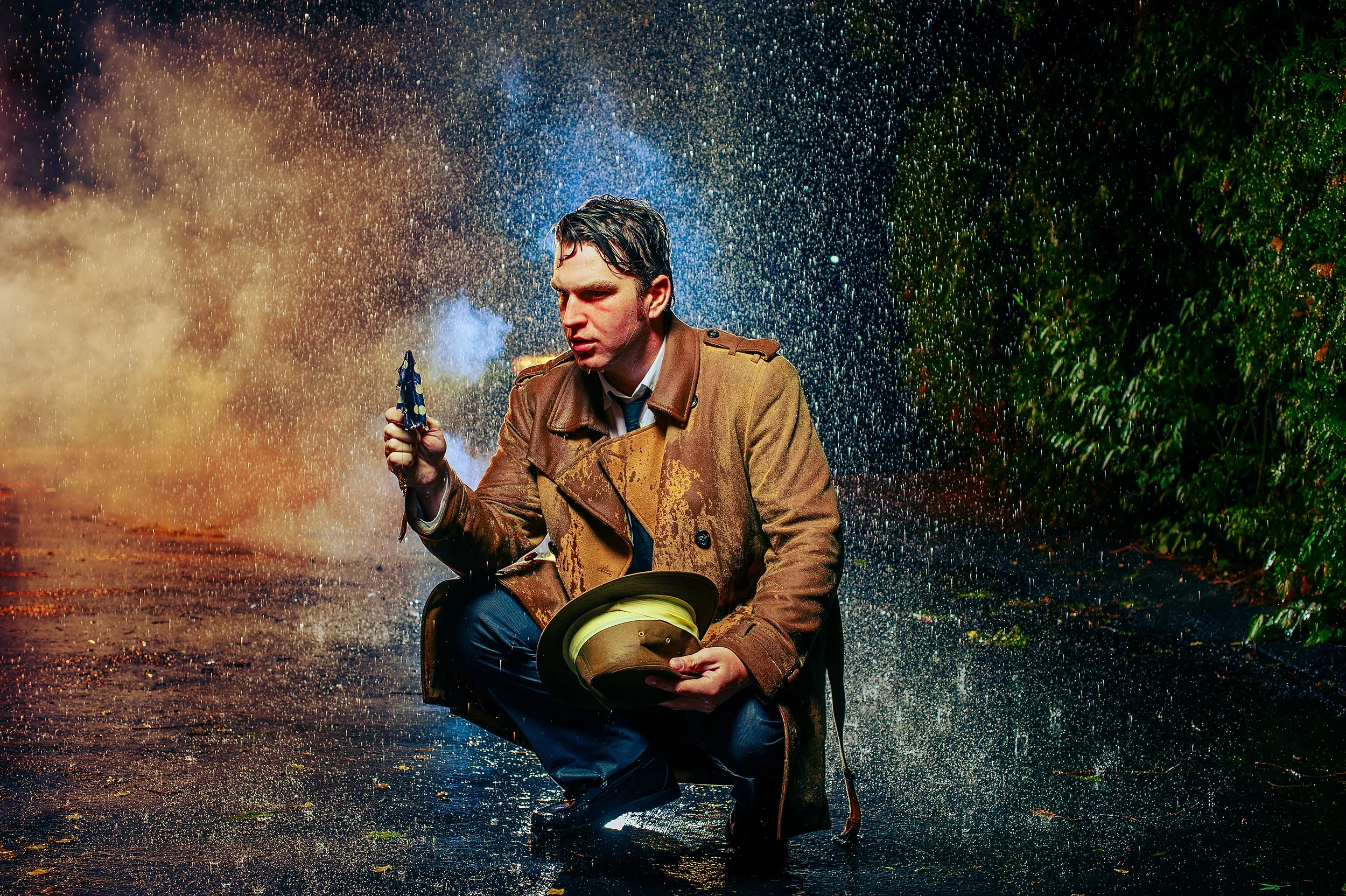A man in a brown trench coat and pouring rain is sitting on a wet street, holding an umbrella and a hat, amidst colorful rain and illuminated night surroundings.