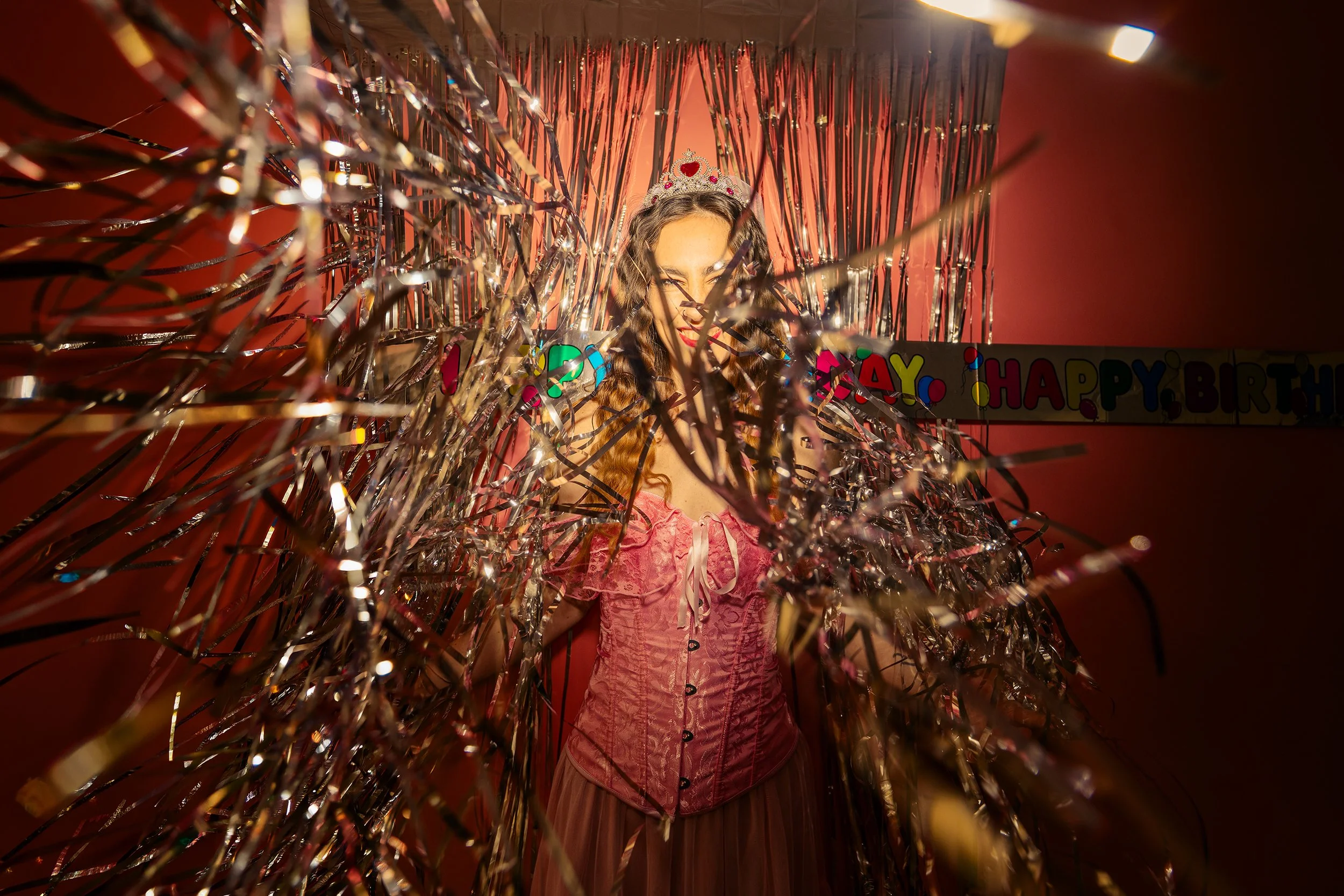 A woman in a pink dress and tiara, with a 'Happy Birthday' banner in the background, is behind gold and silver party streamers.
