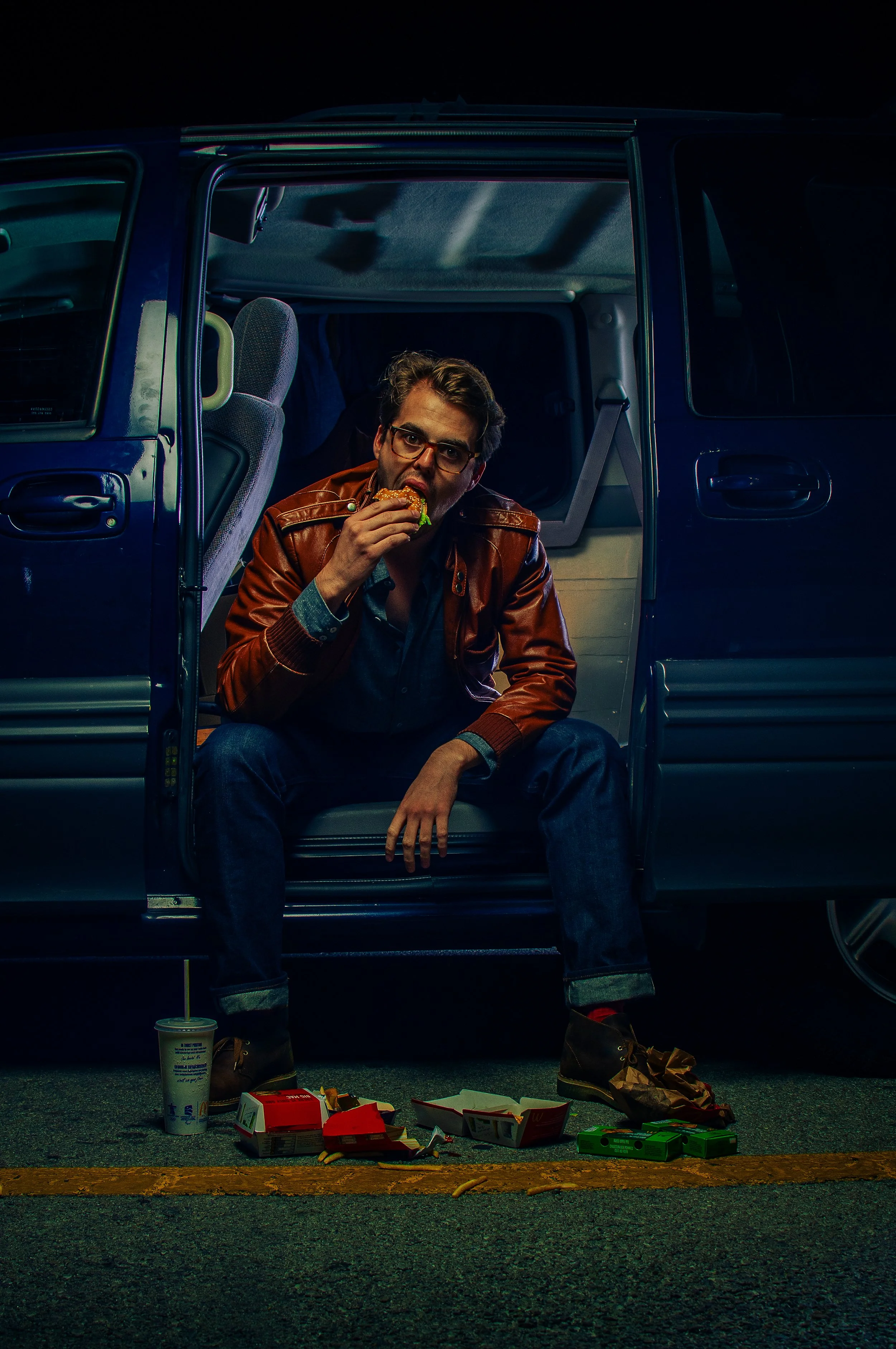 A man with glasses, brown hair, wearing a brown leather jacket and blue jeans, sitting on the edge of an open van at night, eating a burger. scattered fast food containers and soda on the ground in front of him, with a cup with a straw on the left.