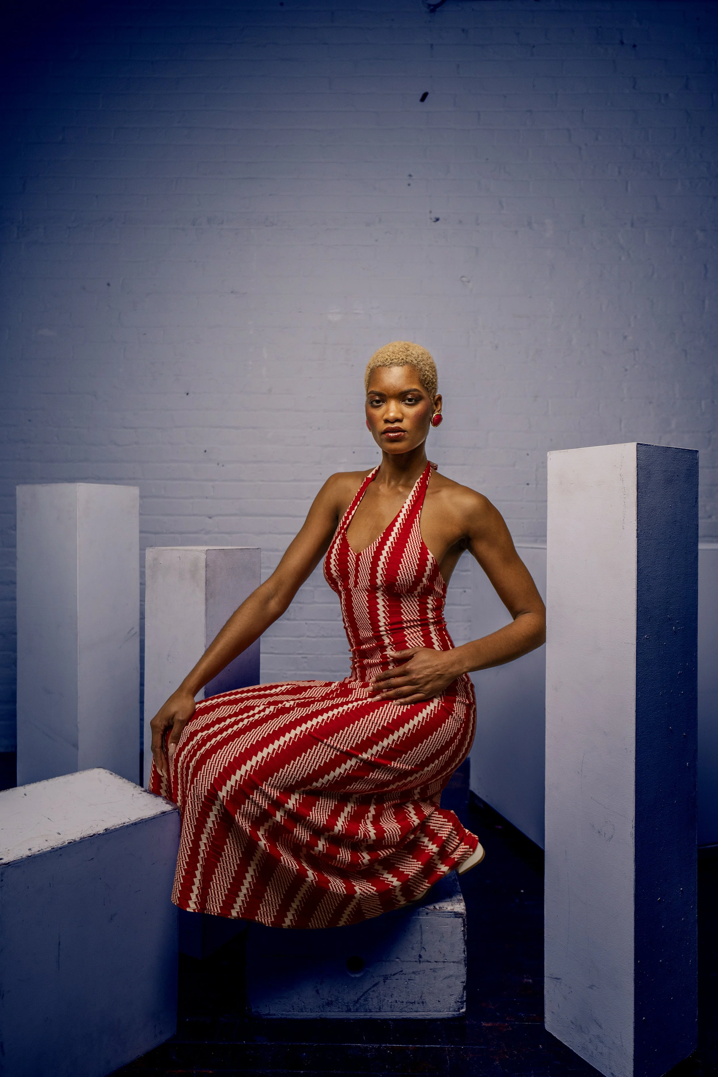 A woman with short blonde hair wearing a red and white patterned halter dress and red earrings, sitting on a white cube, with a white brick wall background and several white and gray cubed structures around her.