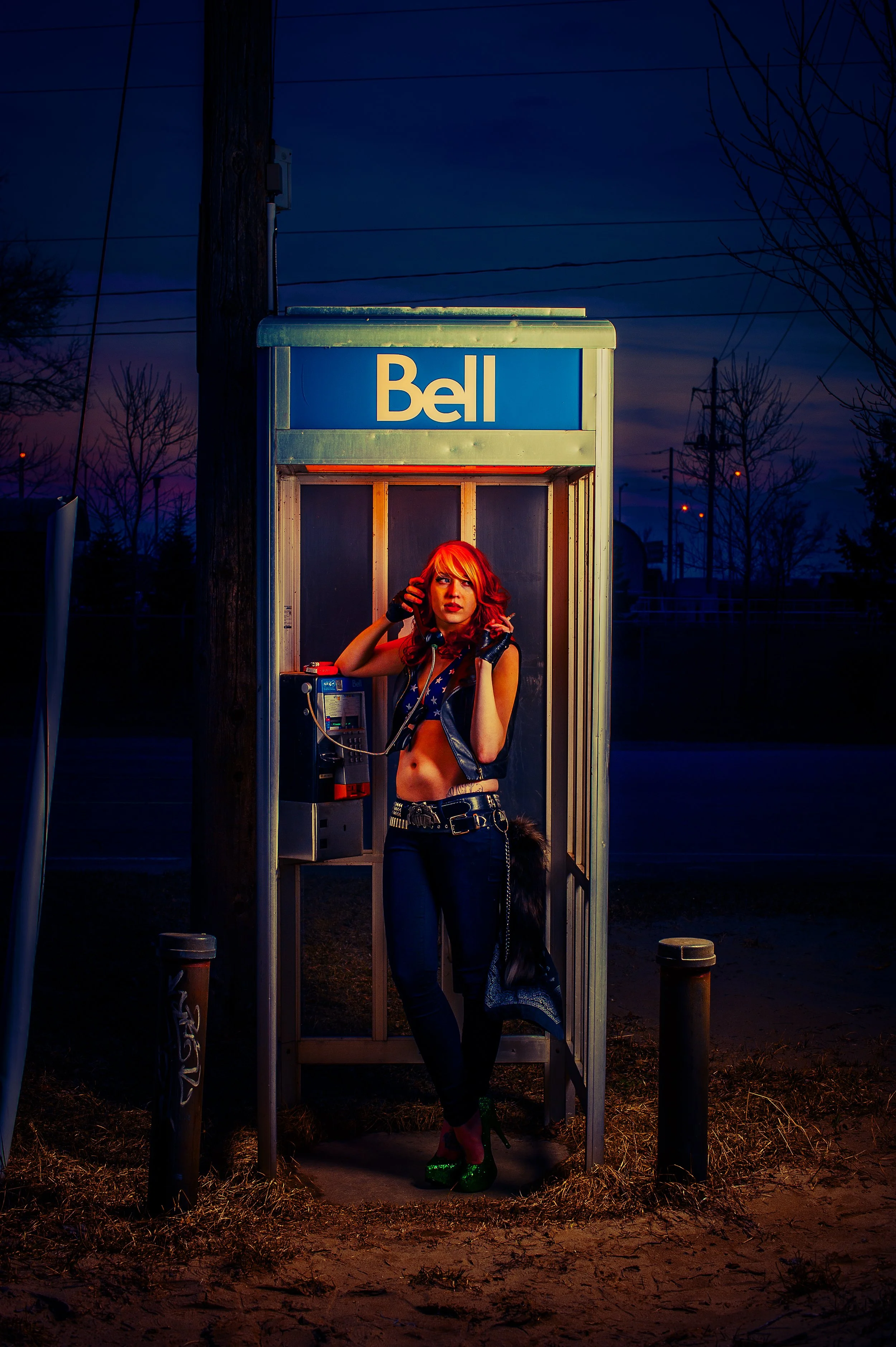A woman with red hair dressed in a dark blue outfit with stars, wearing green platform shoes, standing inside a phone booth at dusk. The booth is branded with a blue and white Bell sign. She is holding the phone receiver to her ear, looking off to th