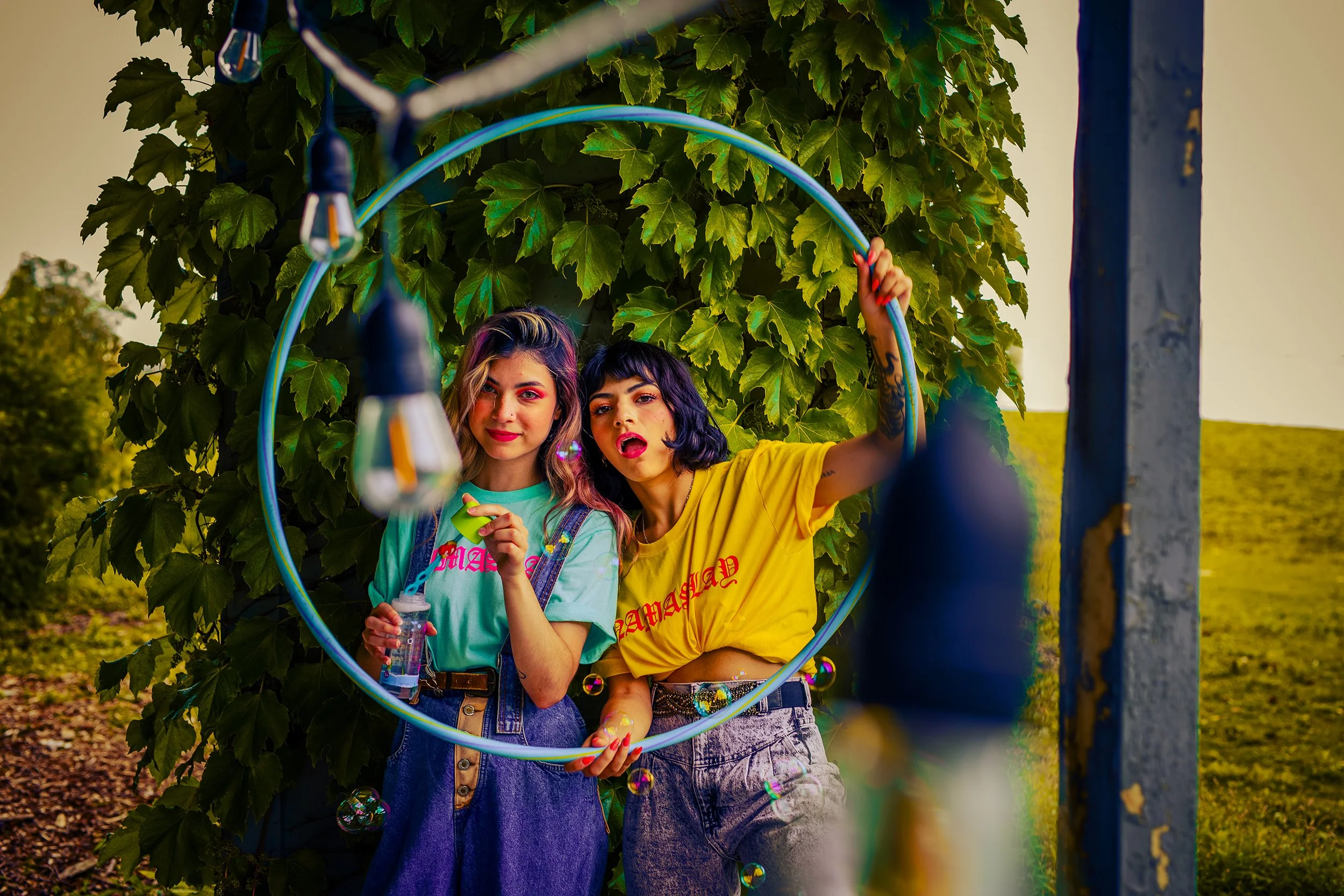 Two young women with colorful hair and makeup playing with bubbles through a hula hoop outdoors near a green leafy bush.