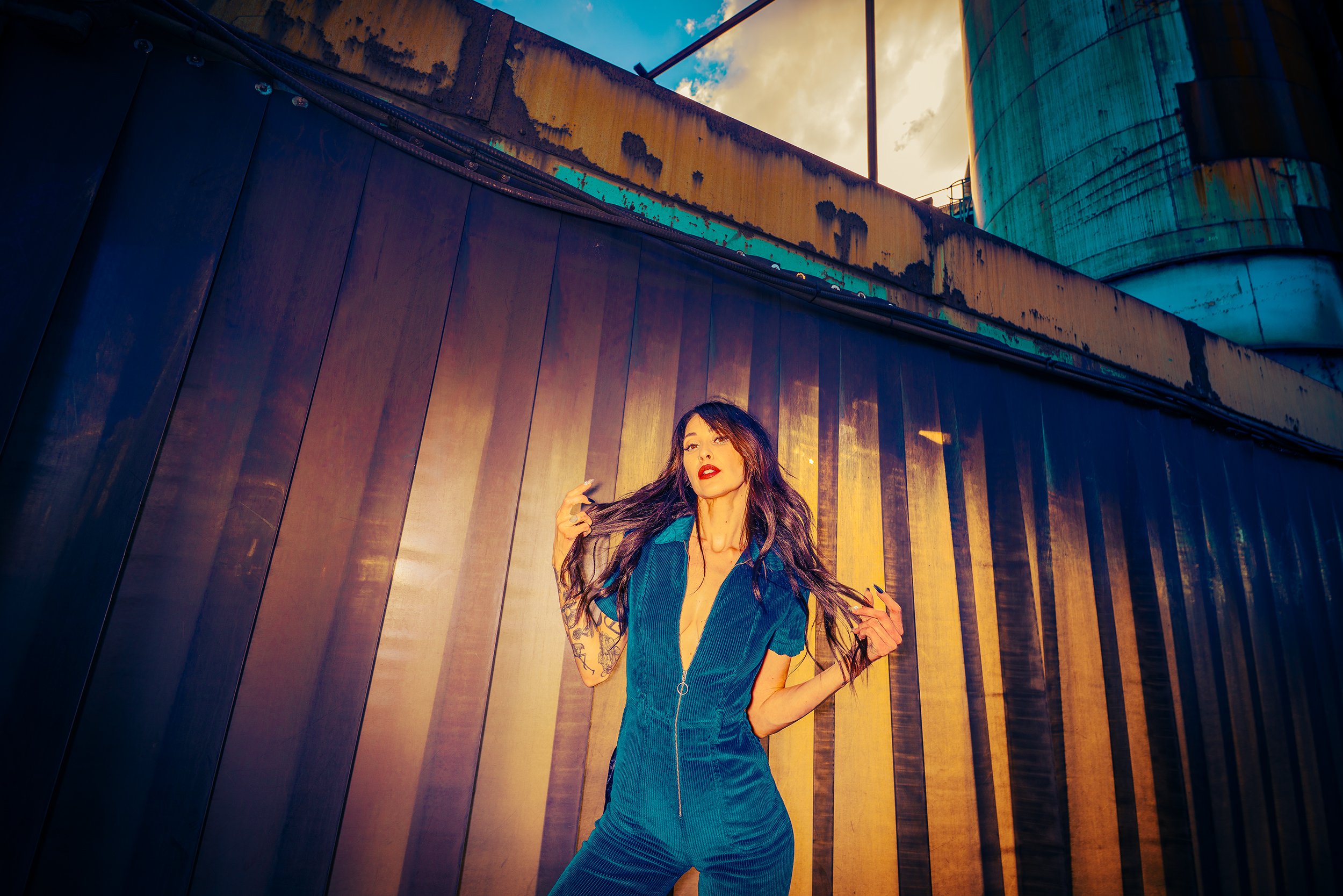 A woman with long dark hair and red lipstick standing against an industrial background of rusted metal and large cylindrical tanks, wearing a dark jacket and blue outfit.