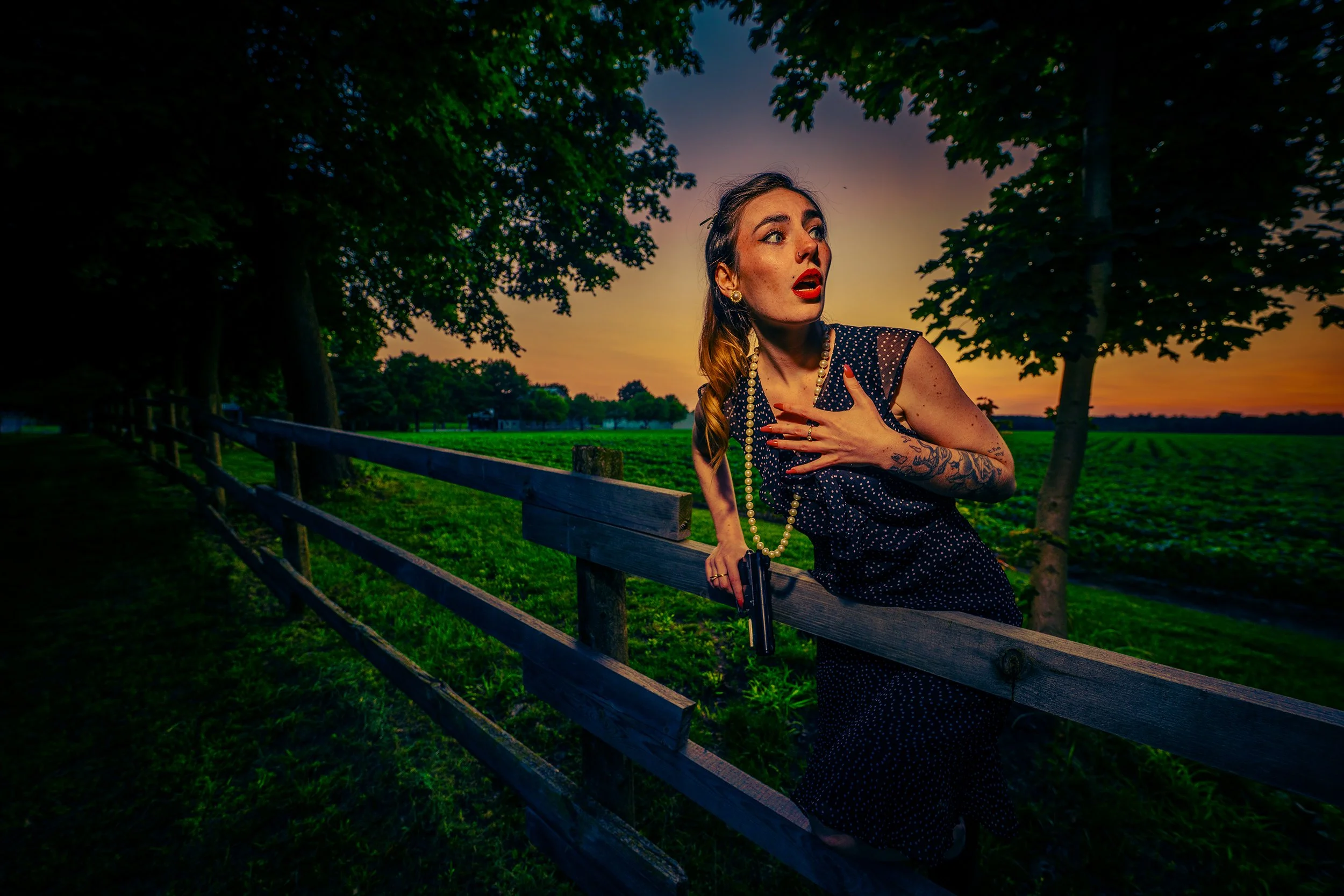 A woman with tattoos and wearing a pearl necklace has her hand on her chest and holds a gun in her other hand, standing next to a wooden fence under trees during sunset.