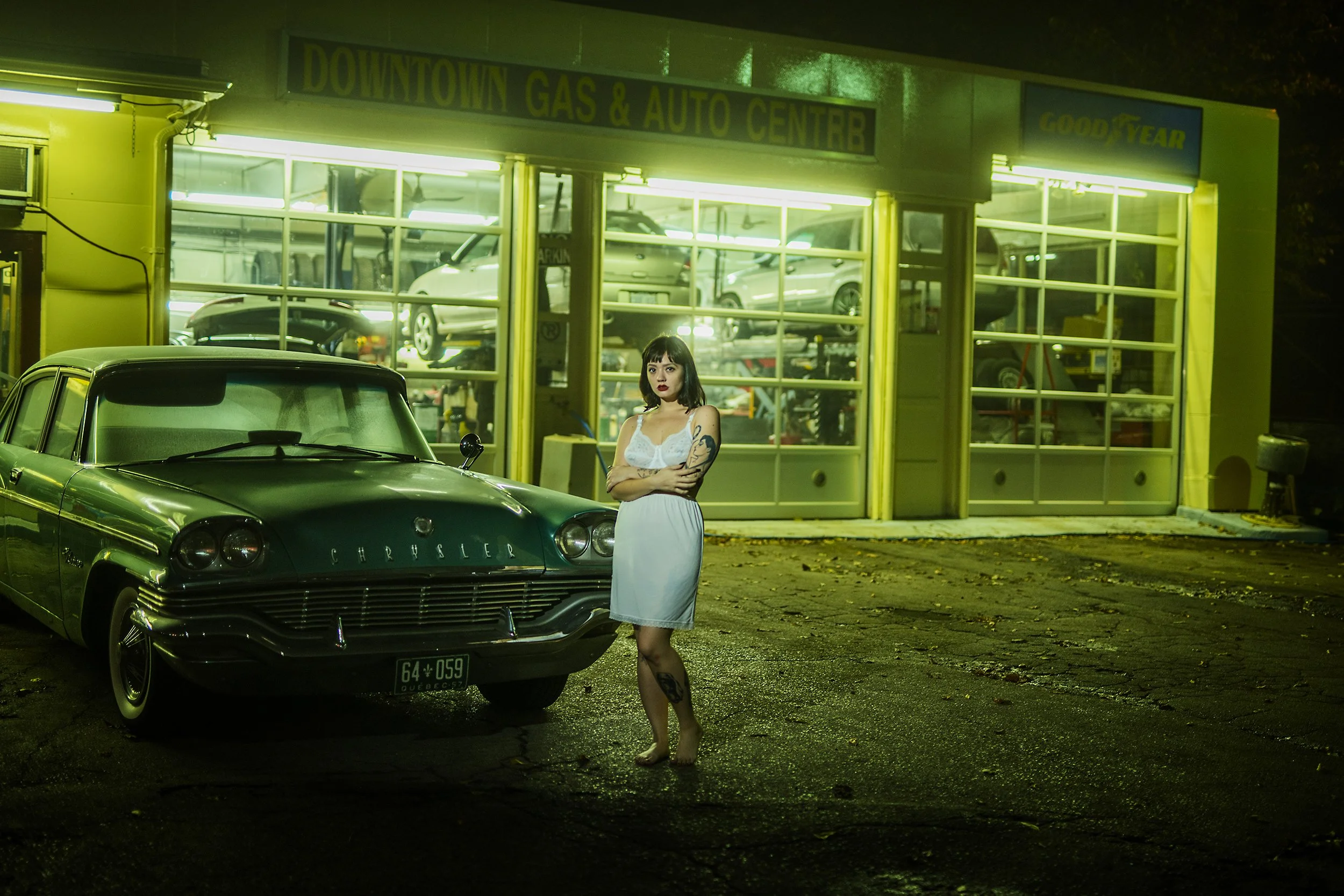 A woman in a white dress standing barefoot in front of a classic green Chrysler car outside a brightly lit auto repair shop at night. The shop has large glass windows displaying cars inside. The woman has tattoos on her arms and legs, dark hair, and 
