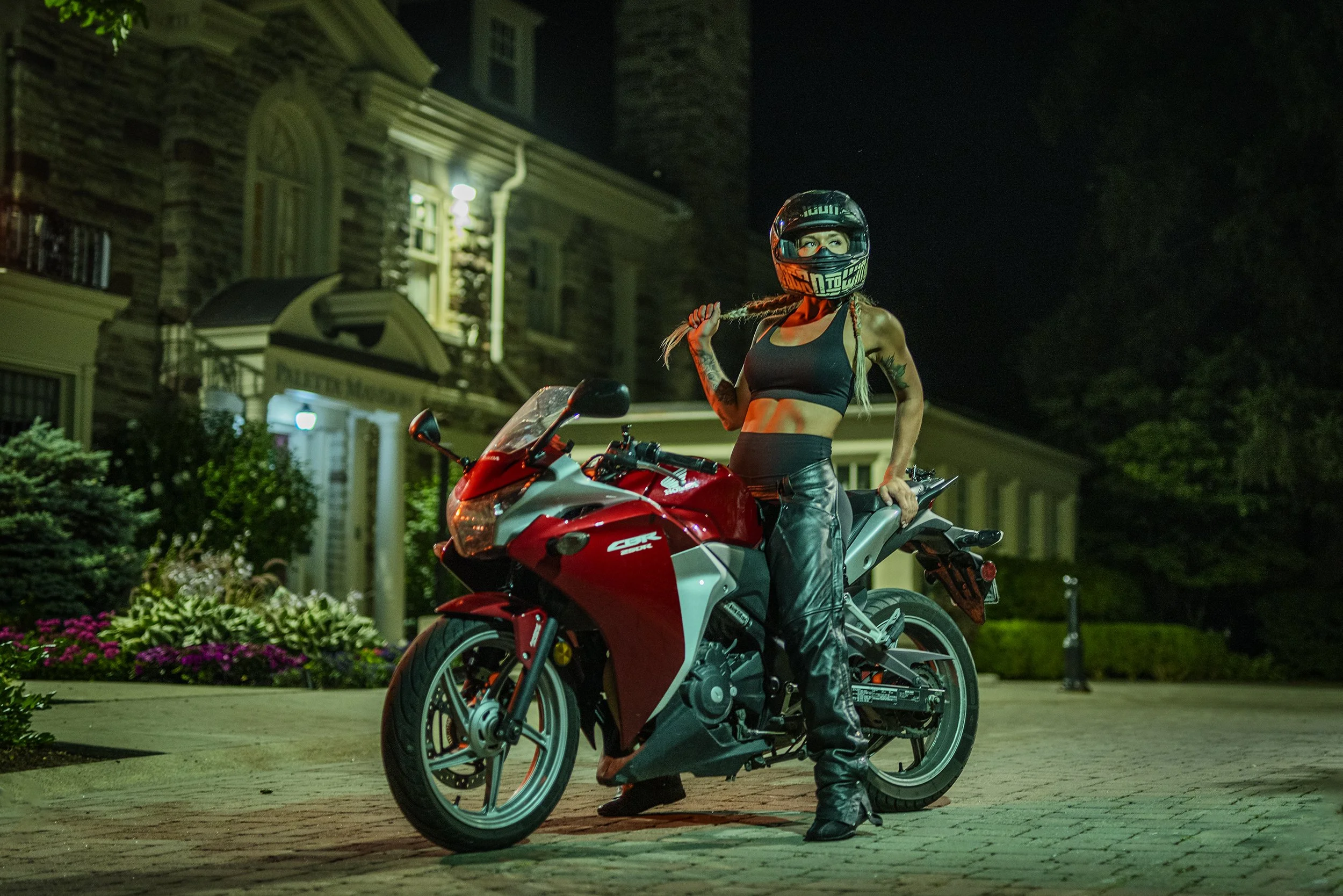 A woman wearing a helmet and black leather pants is standing next to a red and white sport motorcycle on a street at night. She holds a braid of her hair with her right hand. The background includes a brick house with illuminated windows and a garden