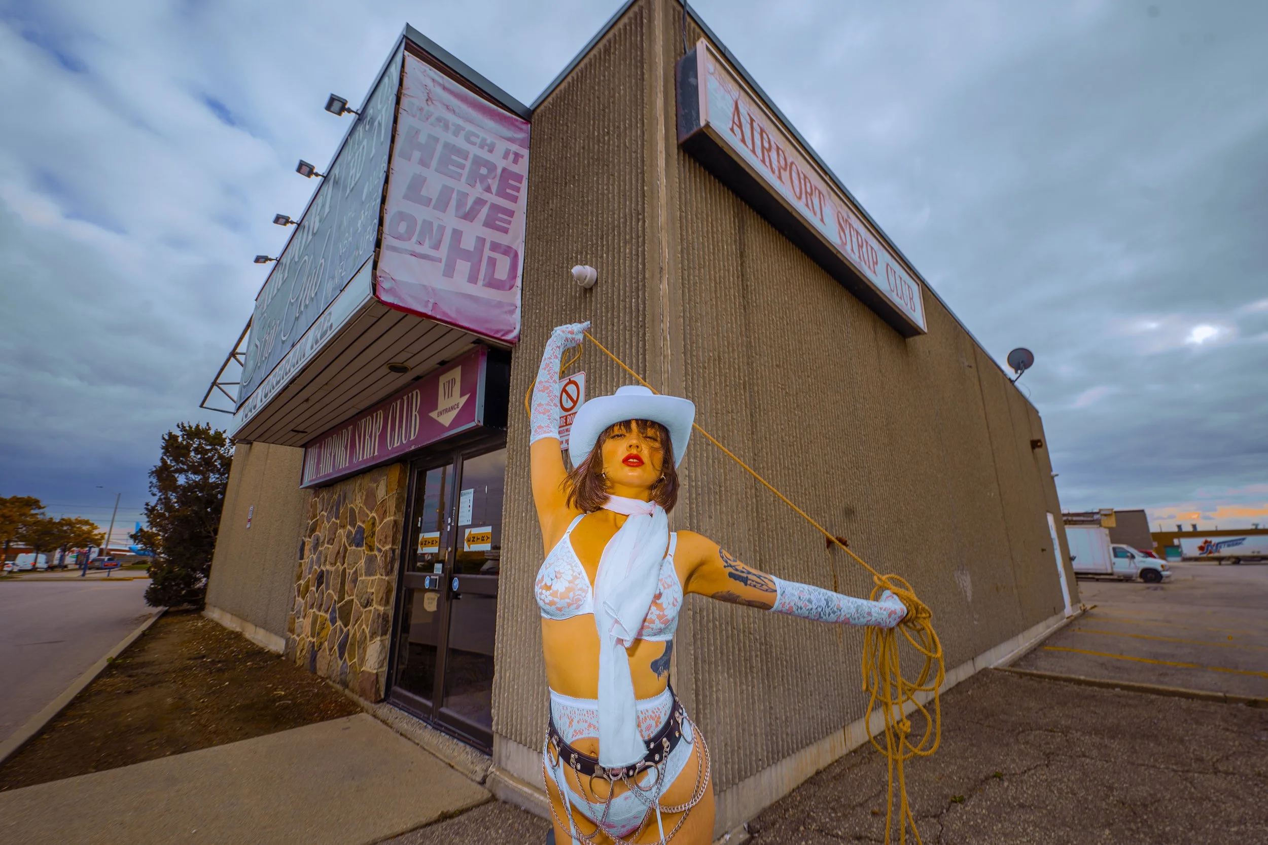 A woman dressed in white lingerie, cowboy hat, and gloves standing outside a building labeled 'Airport Strip Club,' holding a rope and striking a pose. The building has signs for a VIP club and HD streaming, under a cloudy sky.