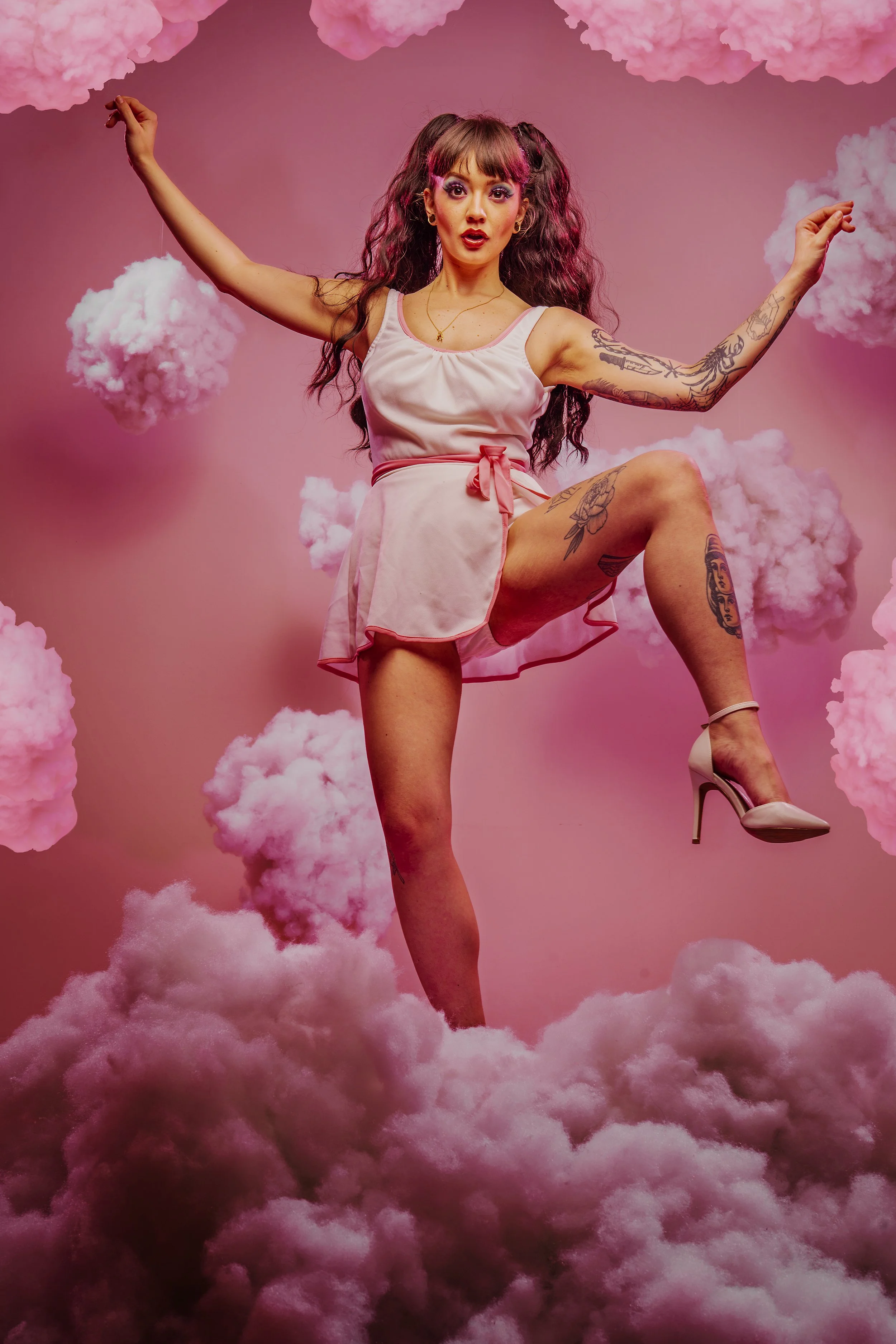 A woman with tattoos standing on pink clouds against a pink background, wearing a silky pink dress and high heels.