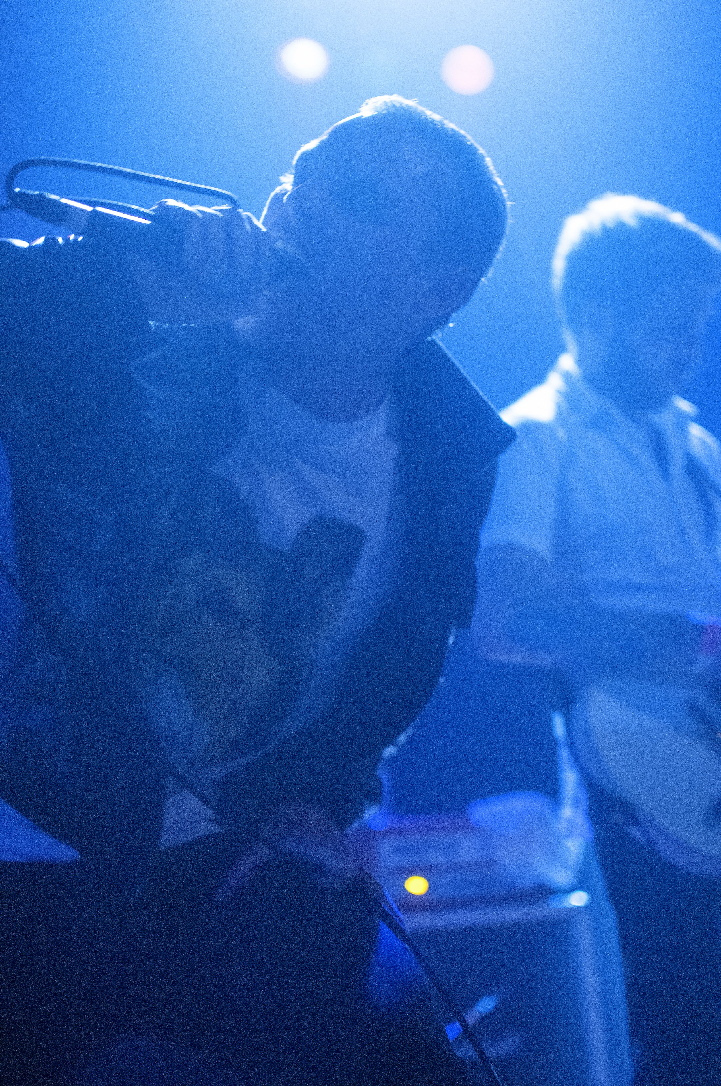 A person holding a microphone close to their mouth with blue stage lighting in the background, and another person partially visible behind them.