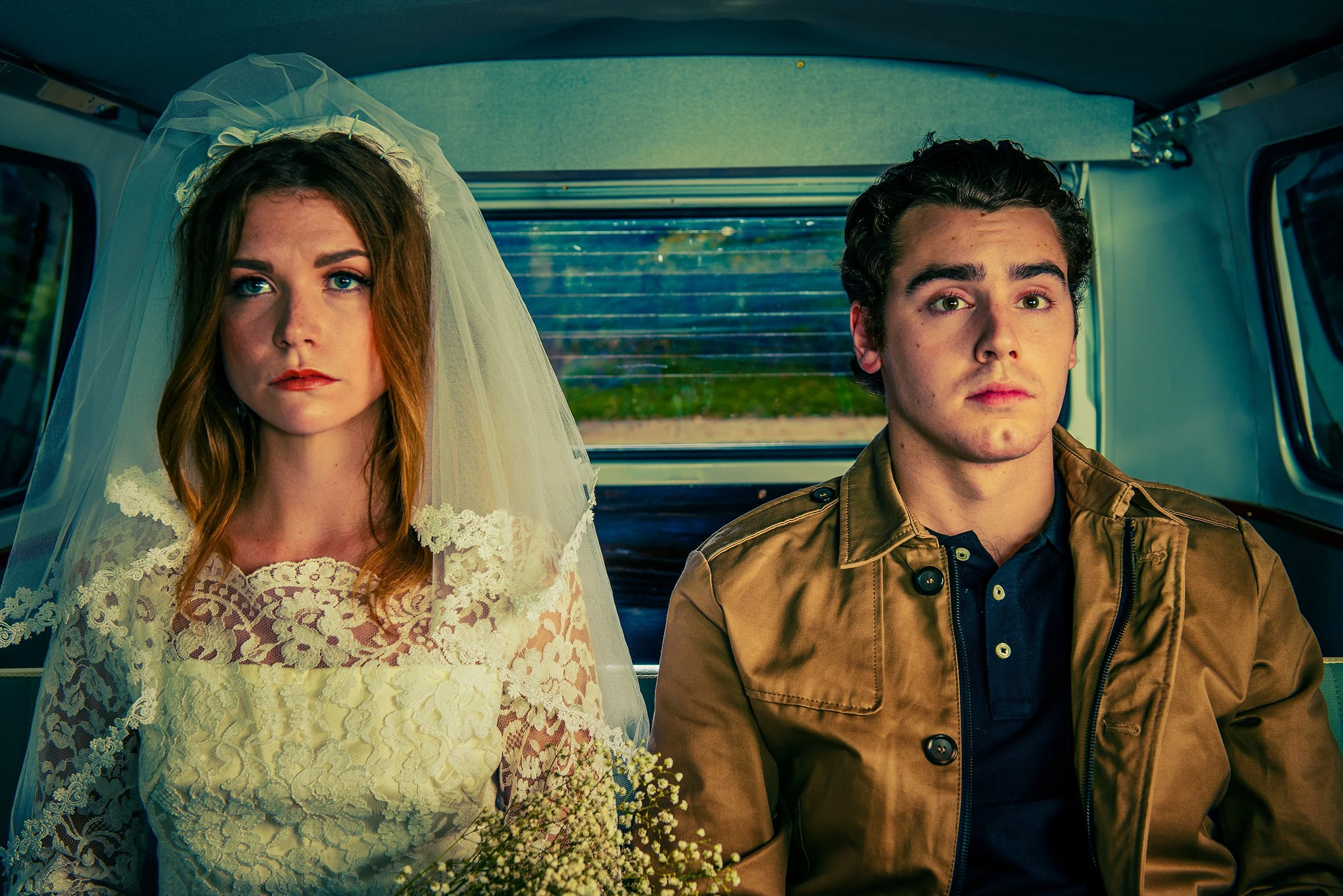 A woman in a wedding dress and veil sitting next to a man in a brown jacket on a bus, both looking at the camera.