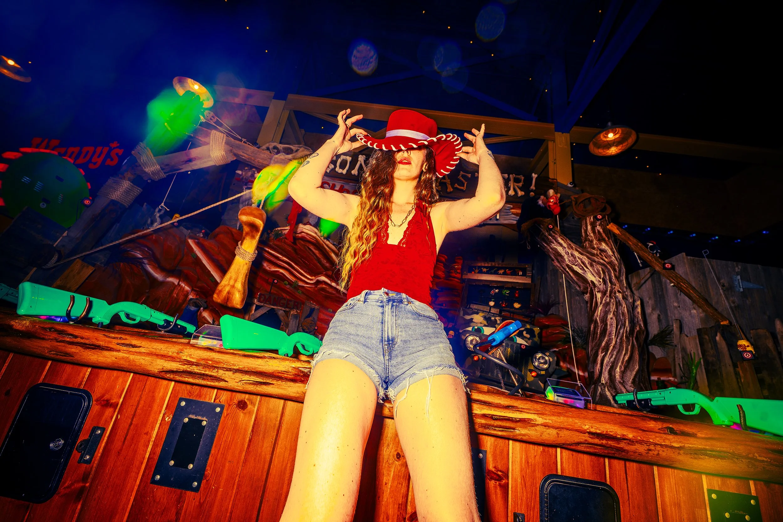 A woman with long curly hair wearing a red dress and a large red and white hat, standing inside a themed arcade or amusement area with various toys, toys guns, and decorative elements around. She is holding her hat with both hands and looking slightl
