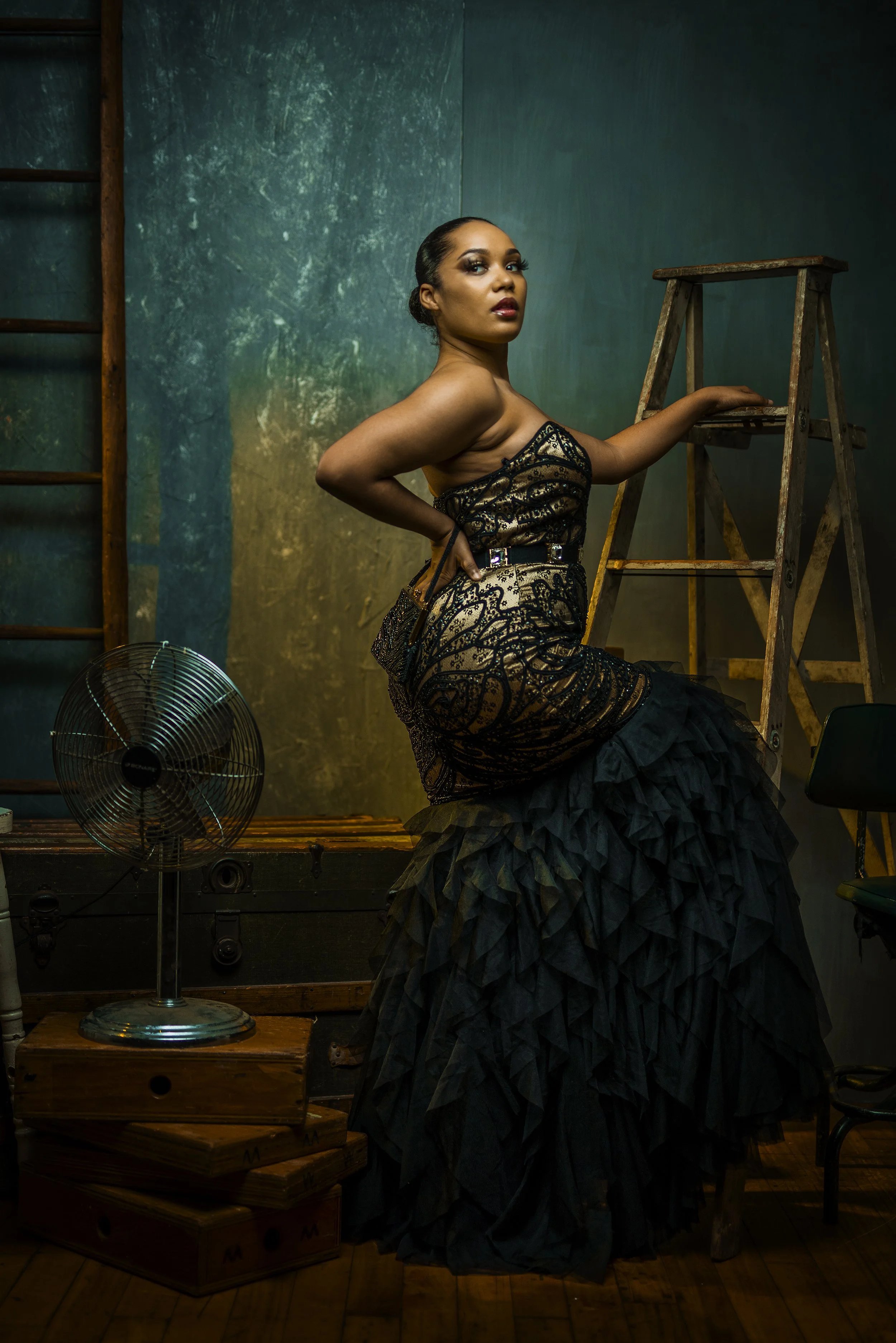 A woman with dark hair styled in an elegant updo, wearing a dark, patterned dress with a ruffled skirt, stands in a room with green and gold textured walls. She is holding onto a wooden ladder with one hand, while her other hand rests on her hip. A v