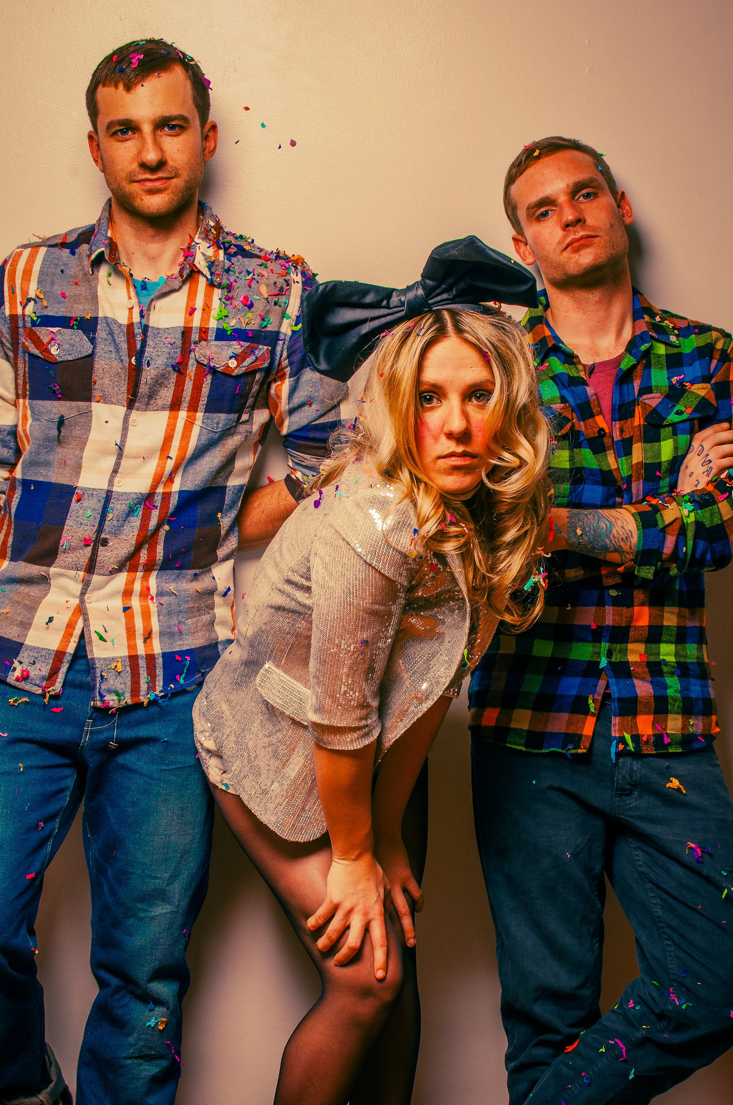 Three people celebrating with colorful confetti falling around them. One woman in the center wearing a headband with a large bow, a shiny top, and black stockings, poses with an intense expression. Two men on either side of her wear casual plaid shir