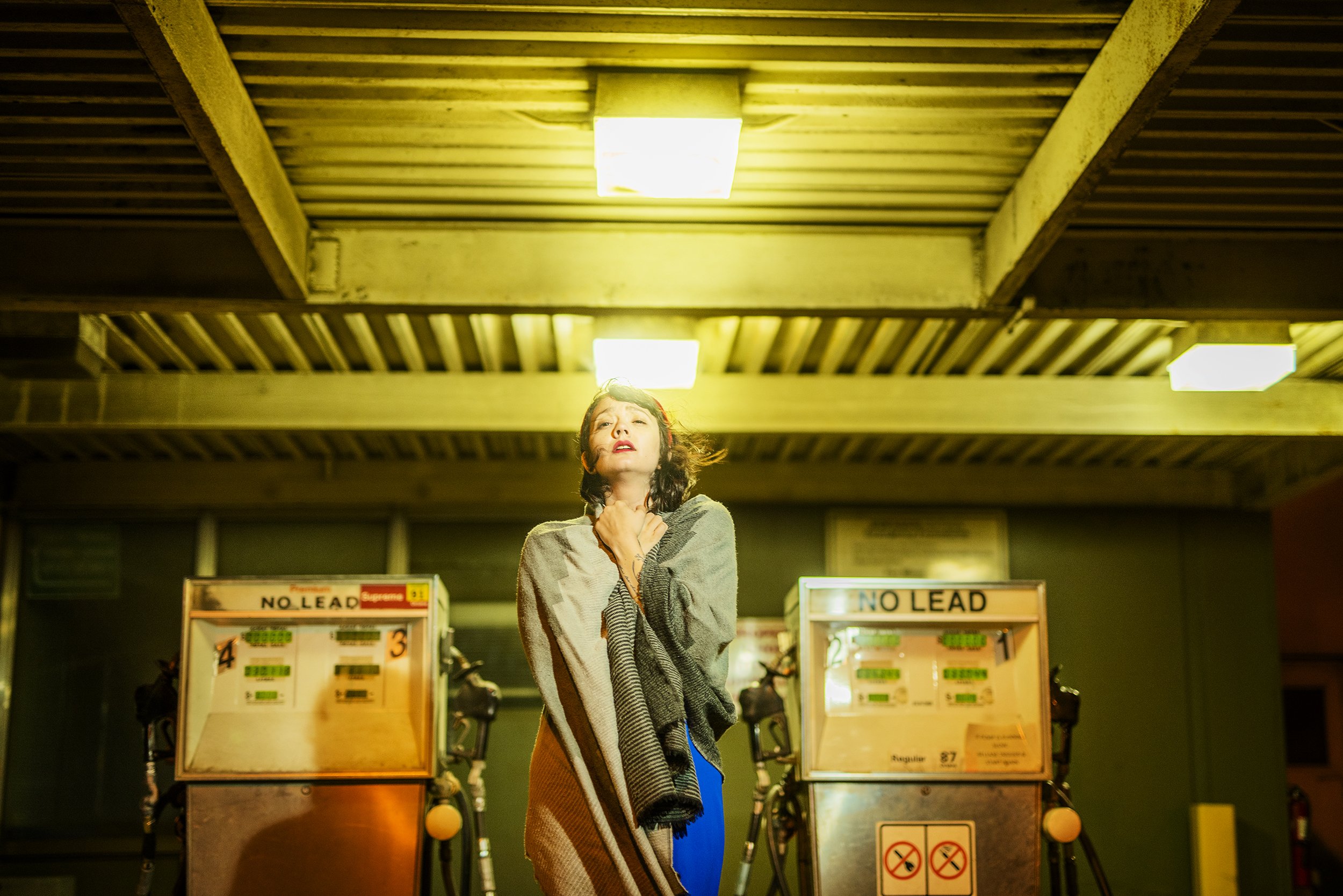 A woman in a striped shawl standing with her hand on her chest inside a parking garage or similar underground setting with warning signs and parking machines behind her.