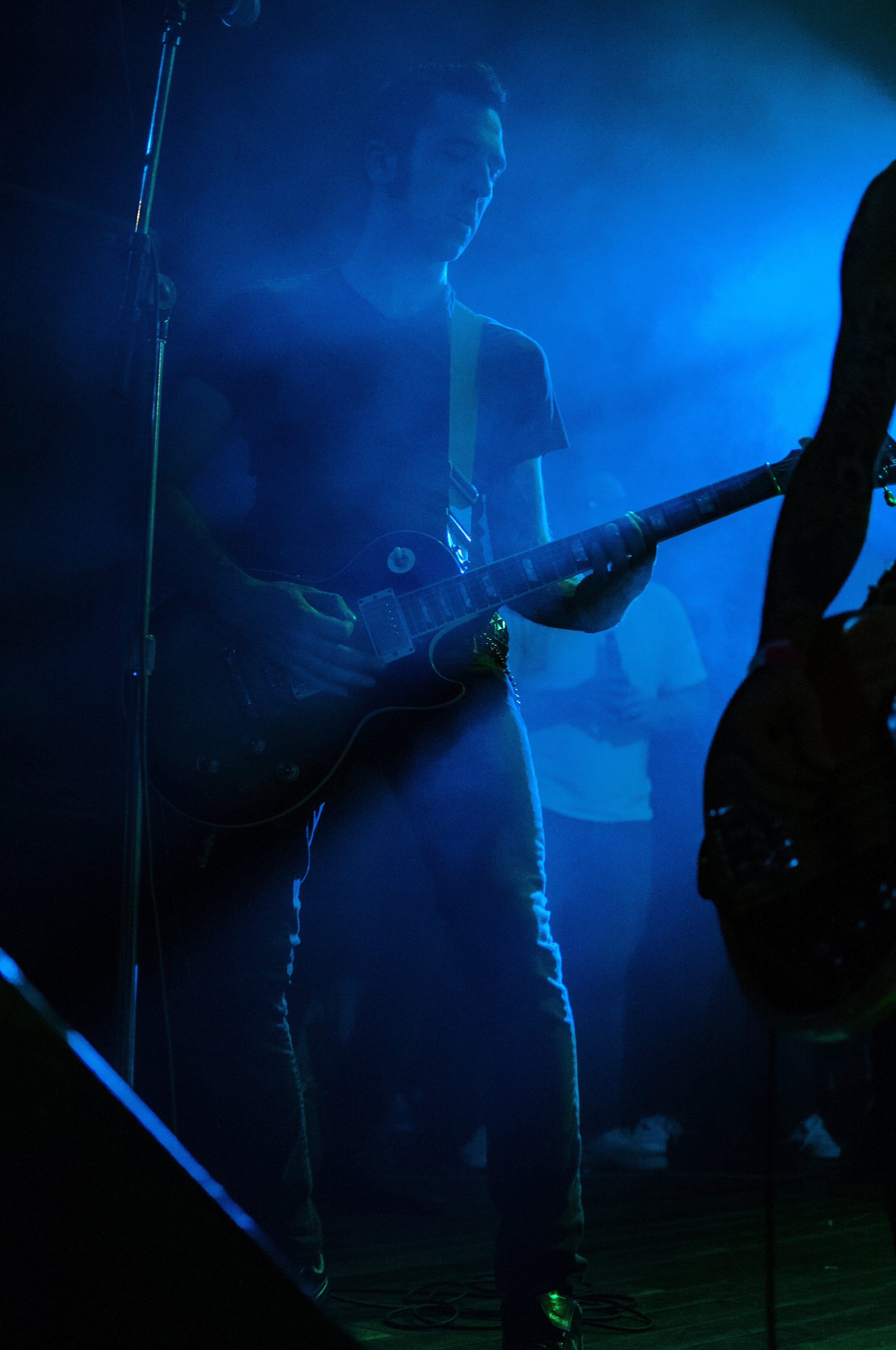 A musician playing an electric guitar on stage with blue stage lighting, person wearing a dark t-shirt and jeans.
