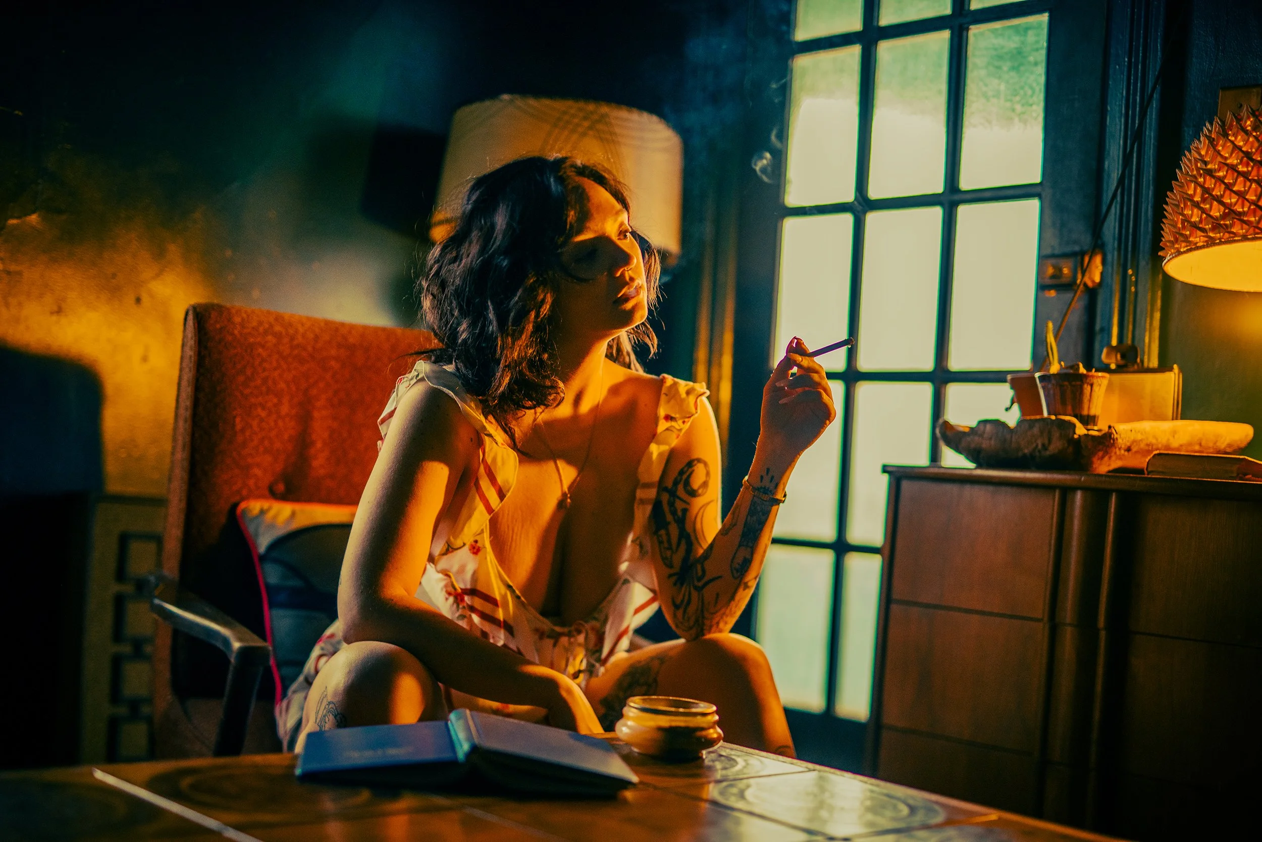 A woman with tattoos and dark wavy hair sitting at a wooden table in a dimly lit room, holding a cigarette near her lips with sunlight streaming through a large window behind her.