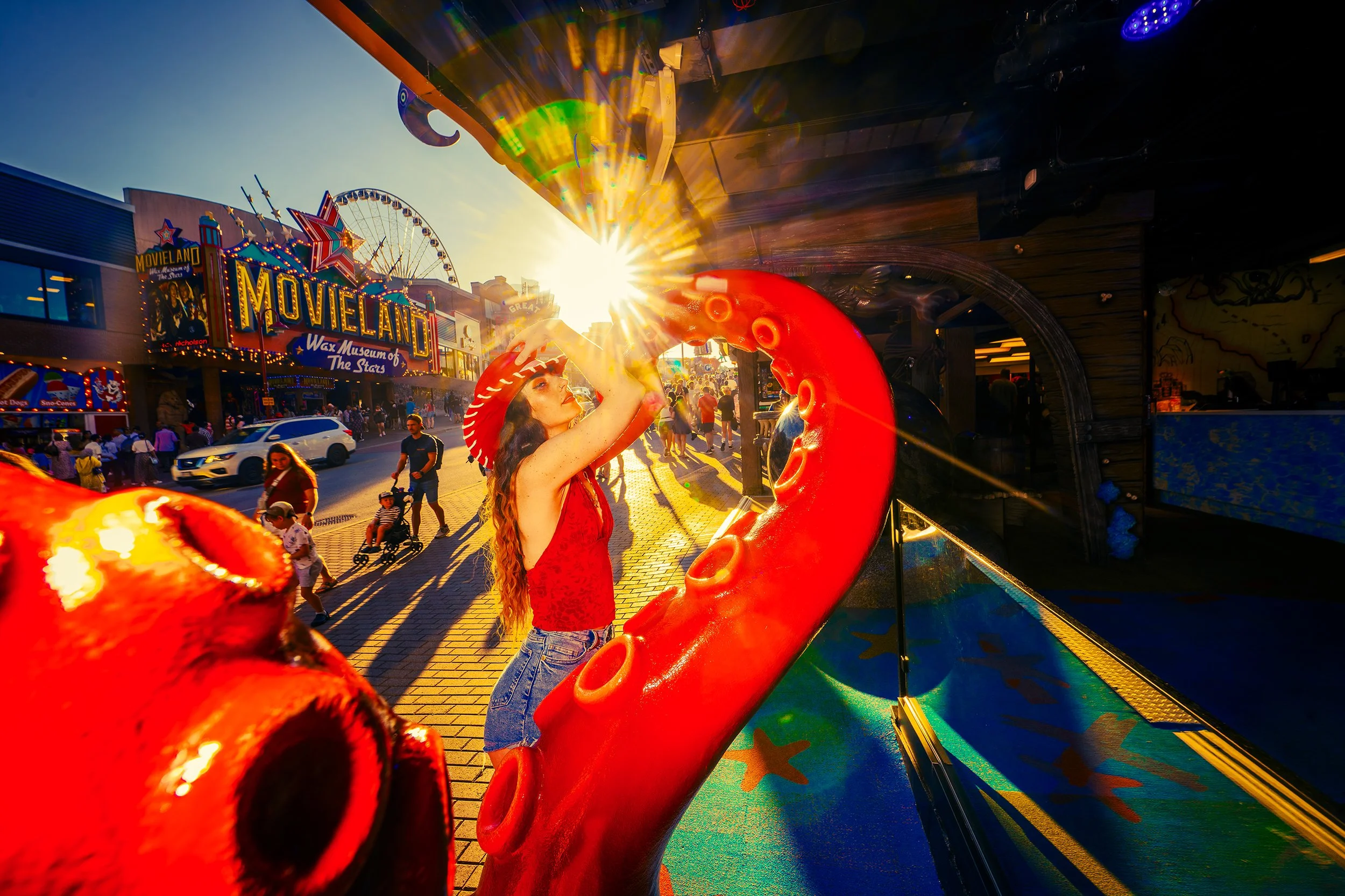 A young woman with long curly hair, wearing a red top and jeans, is standing inside an arcade game at an amusement park at sunset. The scene features bright sunlight, colorful lights, and a crowd of people, with a sign for the movie-themed attraction