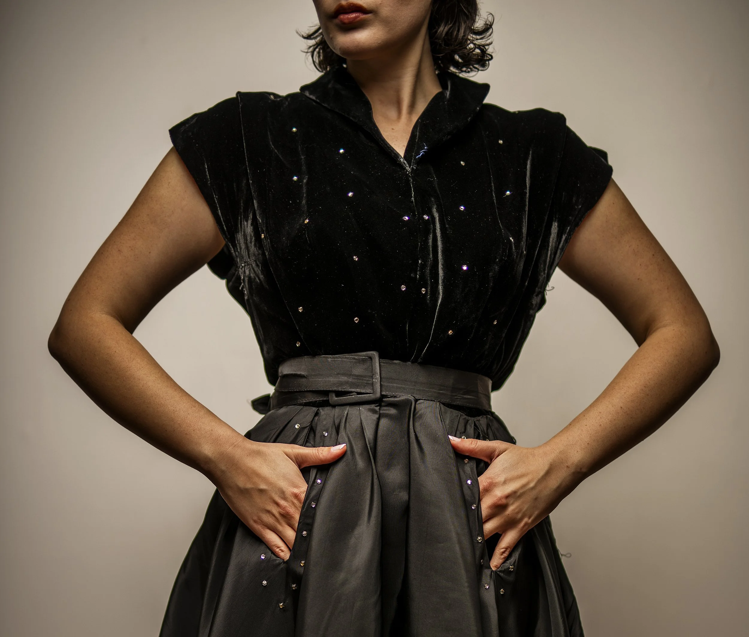 A woman wearing a black sleeveless velvet top with small shiny embellishments and a pleated gray skirt, standing with her hands on her hips in front of a plain gray background.
