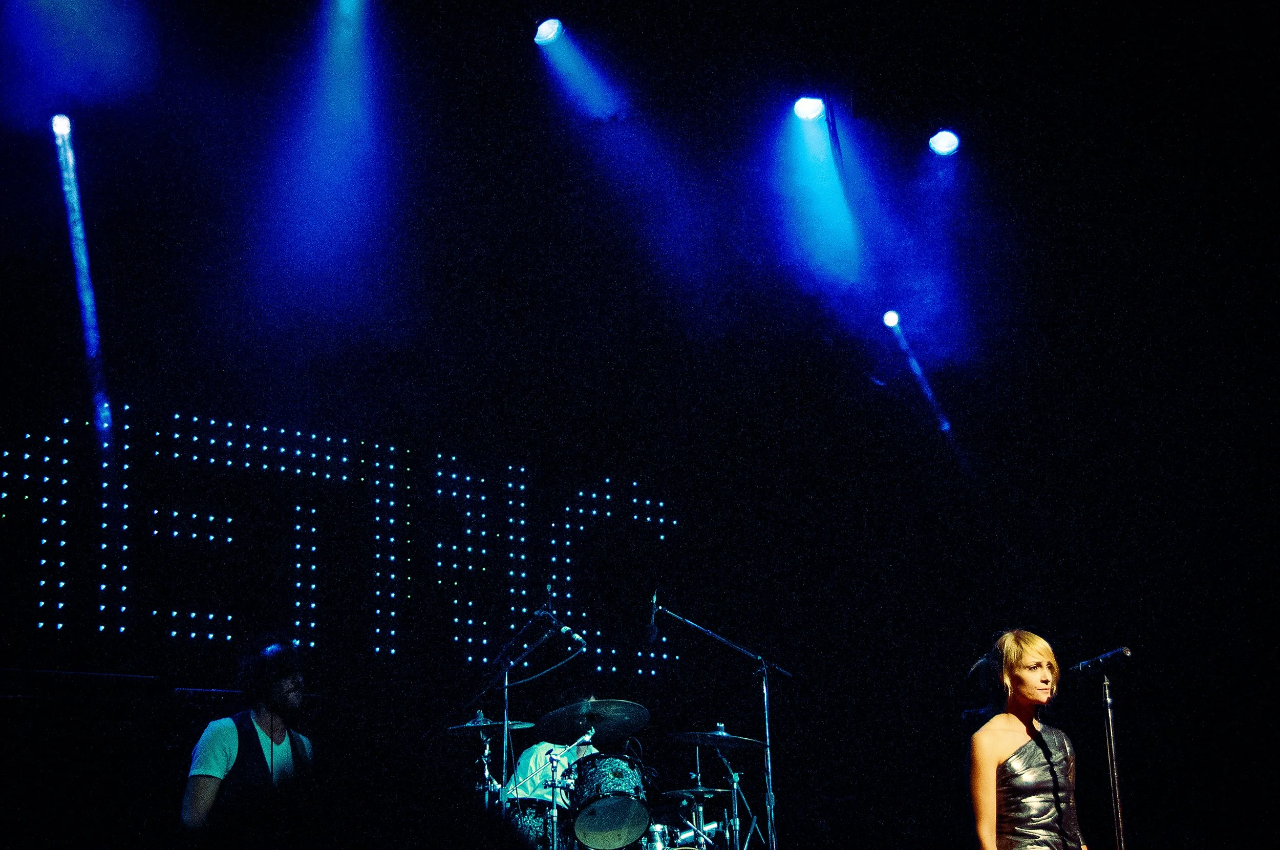 A woman with blonde hair standing on stage in front of a music set, with stage blue lights overhead and a person playing drums in the background.
