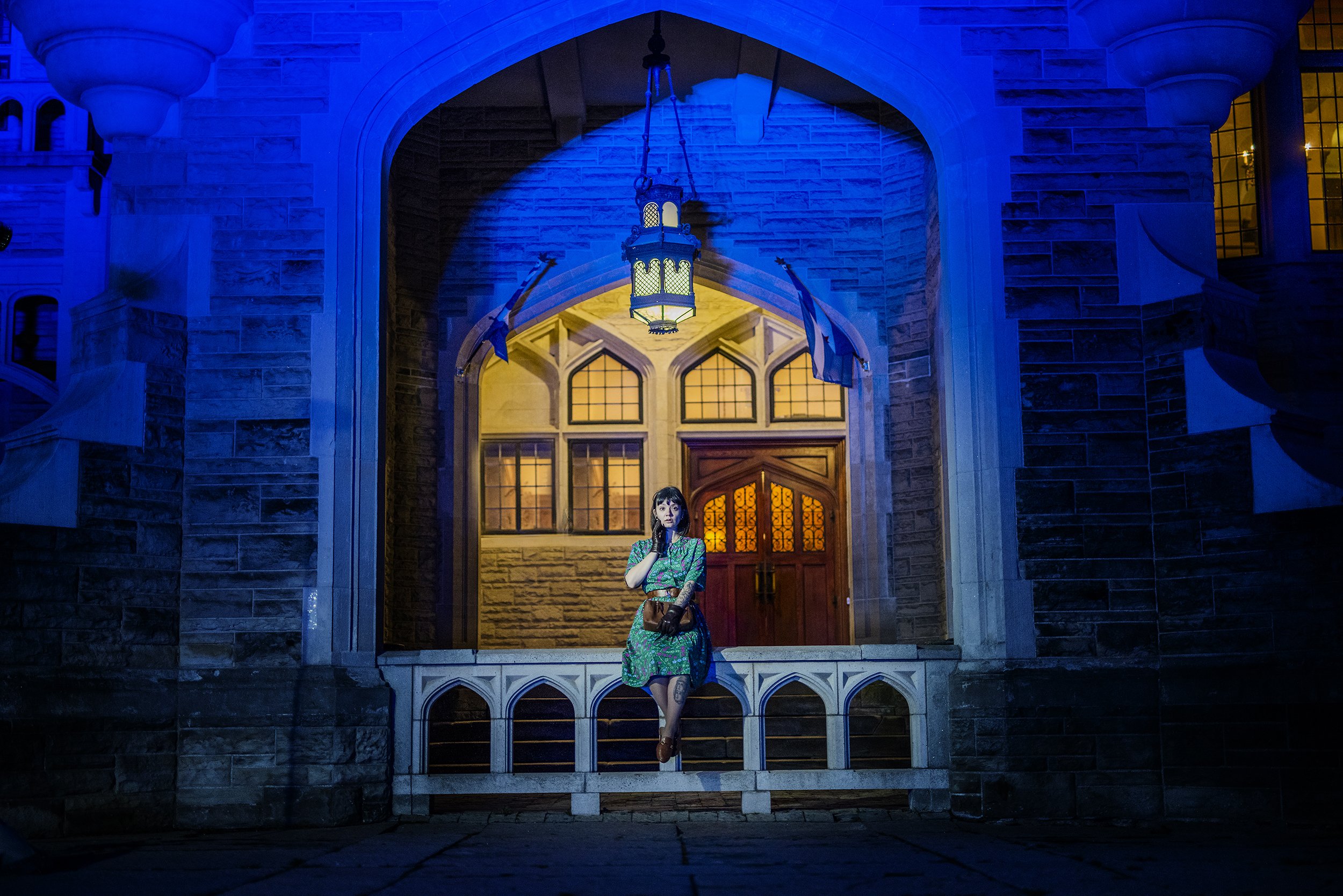 A young woman with dark hair, wearing a green patterned dress and brown boots, sitting on the steps in front of a large, illuminated building with gothic architecture. The scene is lit with blue and warm lighting, creating a dramatic atmosphere.