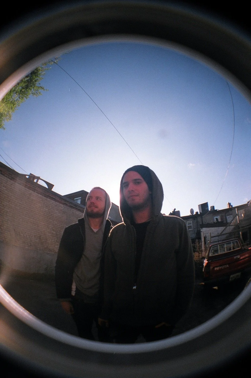 Two men standing outdoors, wearing hooded jackets and hoodies, with a building, parked car, and clear blue sky in the background, seen through a circular fisheye lens.