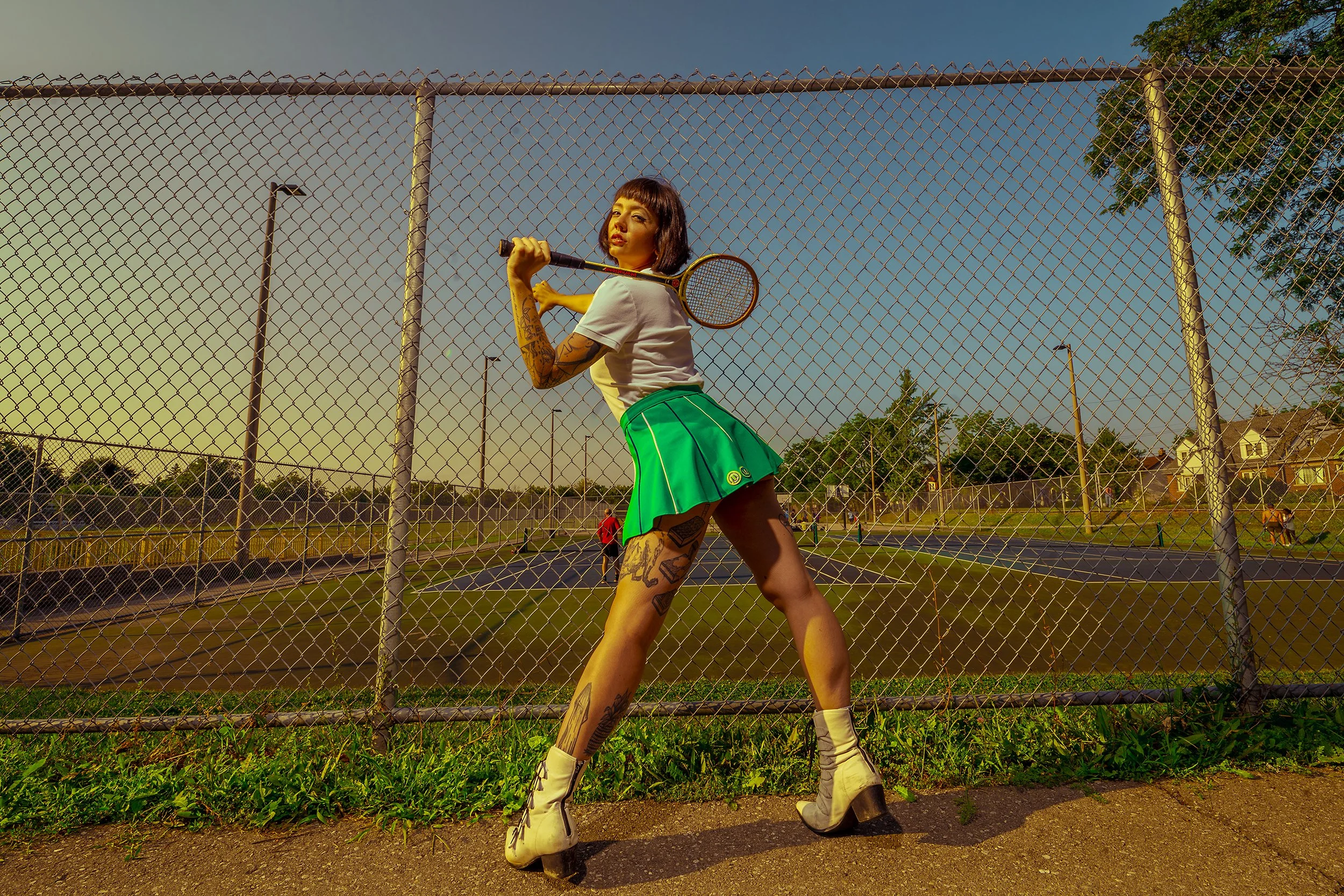 A woman with short black hair, tattoos, and white boots, stands behind a chain-link fence on a tennis court at sunset, holding a tennis racket over her shoulder.