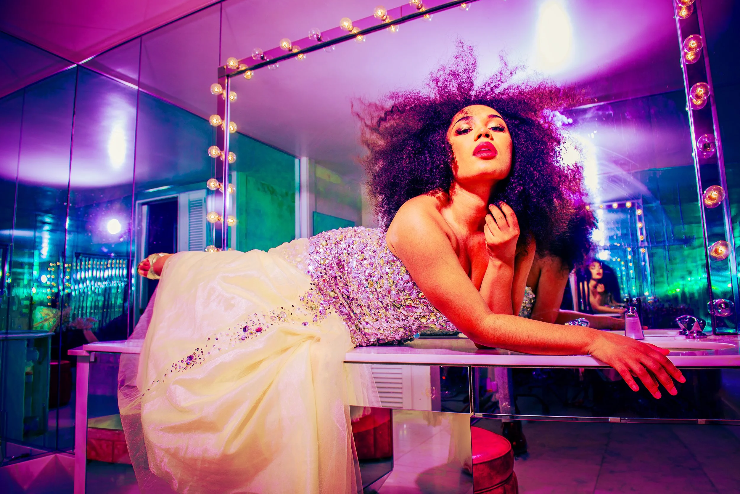 A woman with voluminous curly hair and bold lipstick resting on a vanity table with bright, colorful mirror lights around her, in a vibrant, neon-lit room.