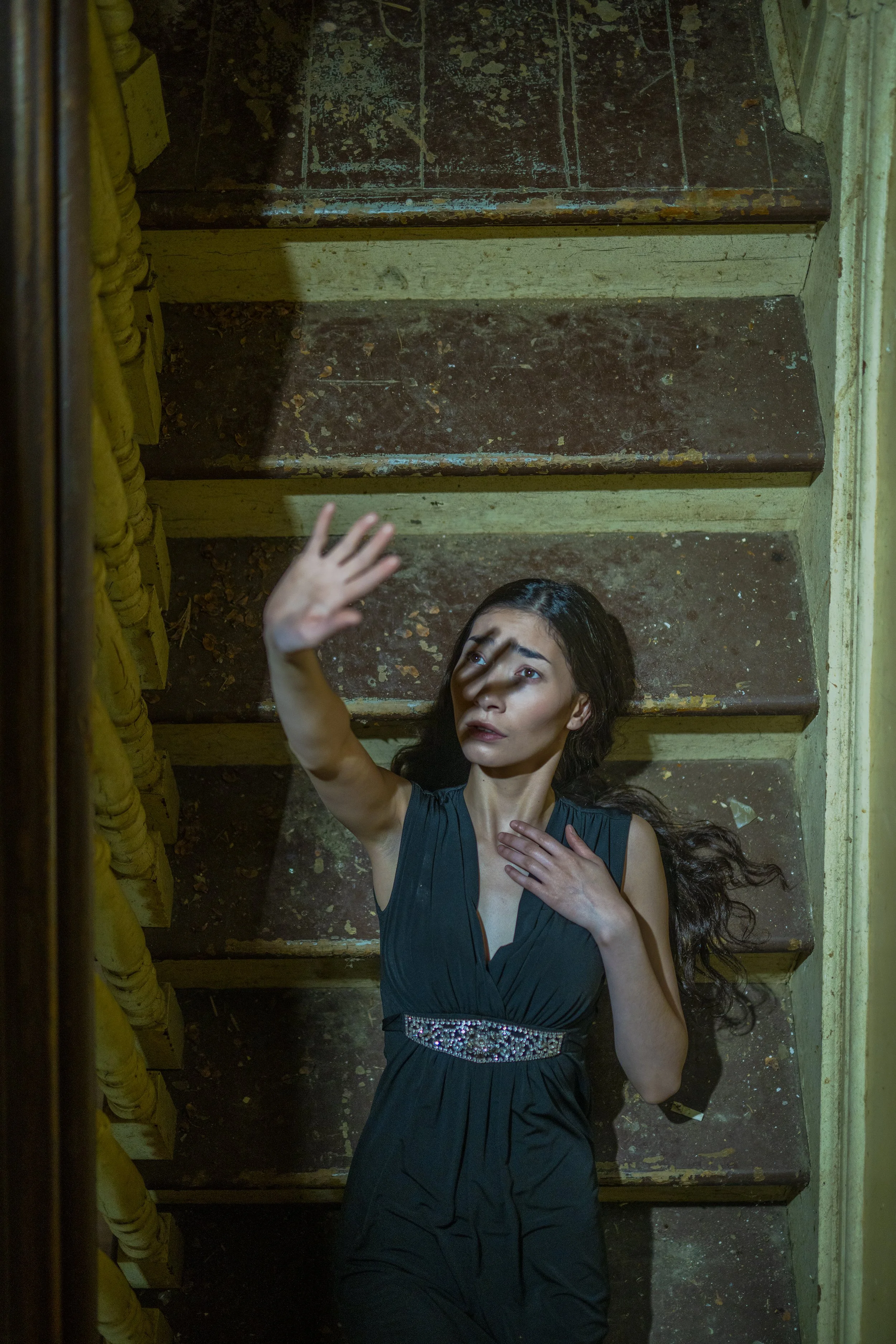 A woman with long dark hair lying on a staircase with yellow sides, wearing a dark teal dress with a sparkling belt. She is looking up and reaching her hand upward, with her other hand on her chest.