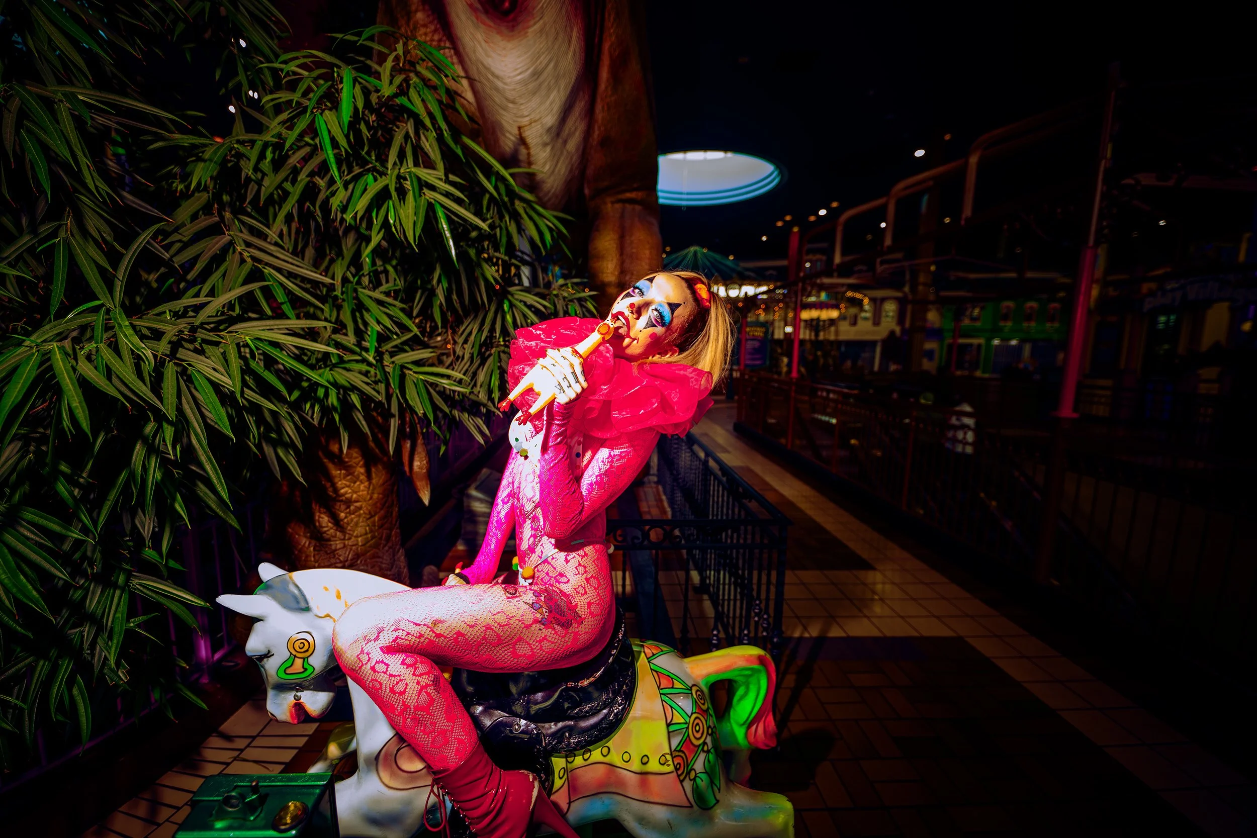 A person dressed as a clown with face paint, wearing a pink outfit, sitting on a colorful carousel horse in an indoor setting with greenery and string lights.