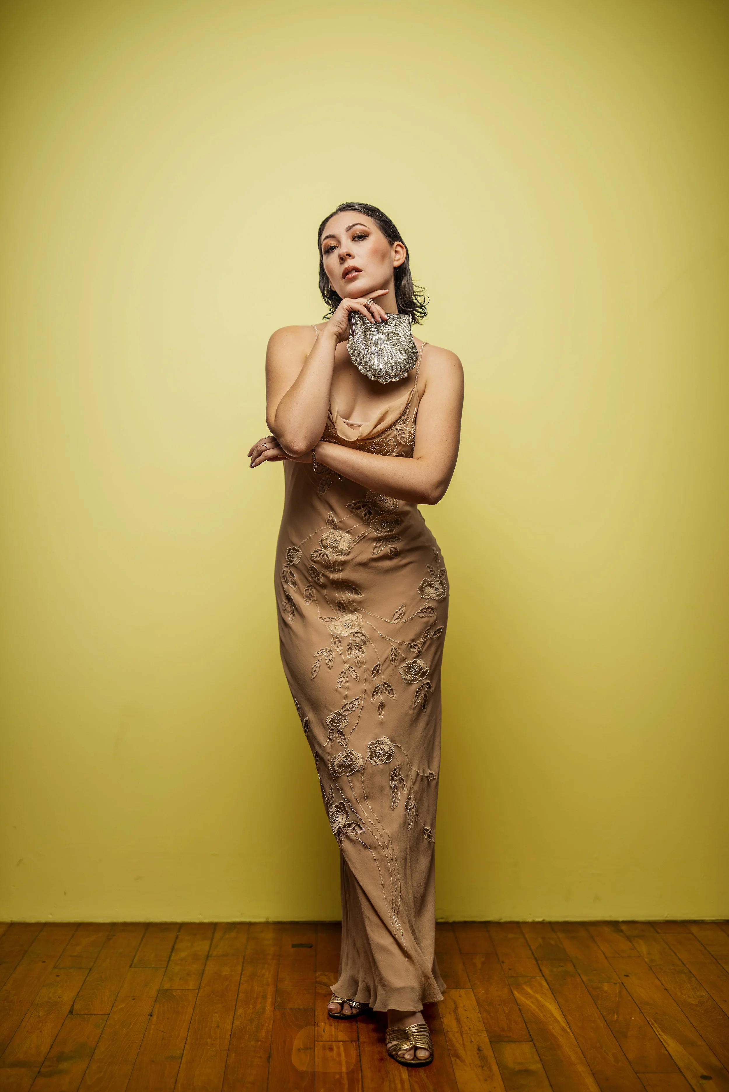 A woman with dark hair and makeup wearing a beige satin gown with embroidered floral designs, holding a silver seashell-shaped clutch, standing against a yellow background.
