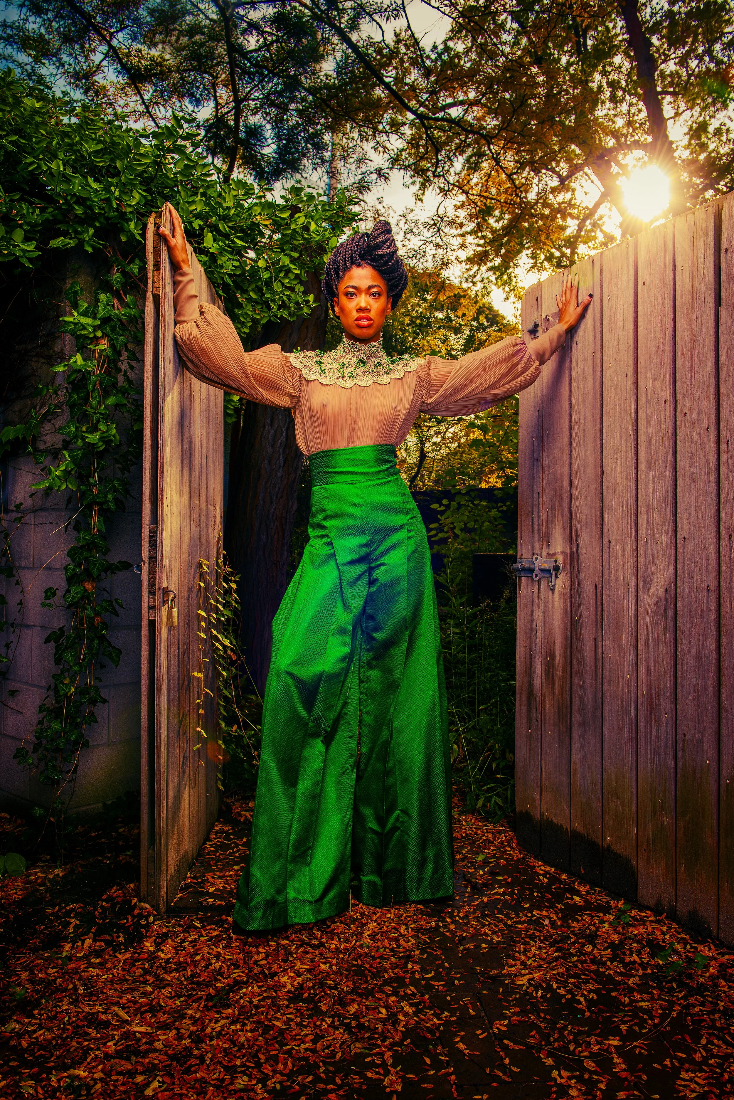 A woman wearing a green skirt and a beige vintage blouse with lace details stands behind a wooden gate in an outdoor setting with trees and autumn leaves on the ground. The sun is setting or rising, casting a warm glow.